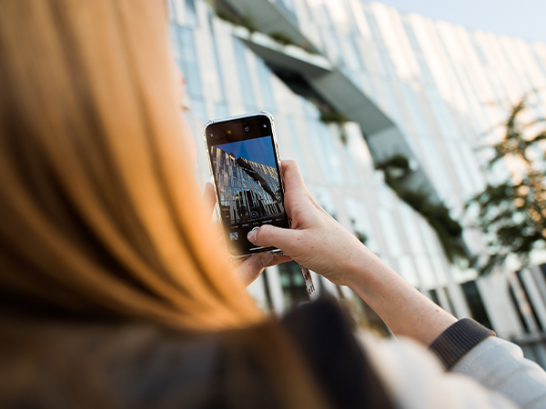 Eine junge rothaarige Frau macht mit ihrem Smartphone ein Foto von einem modernen Gebäude mit Glaswand.