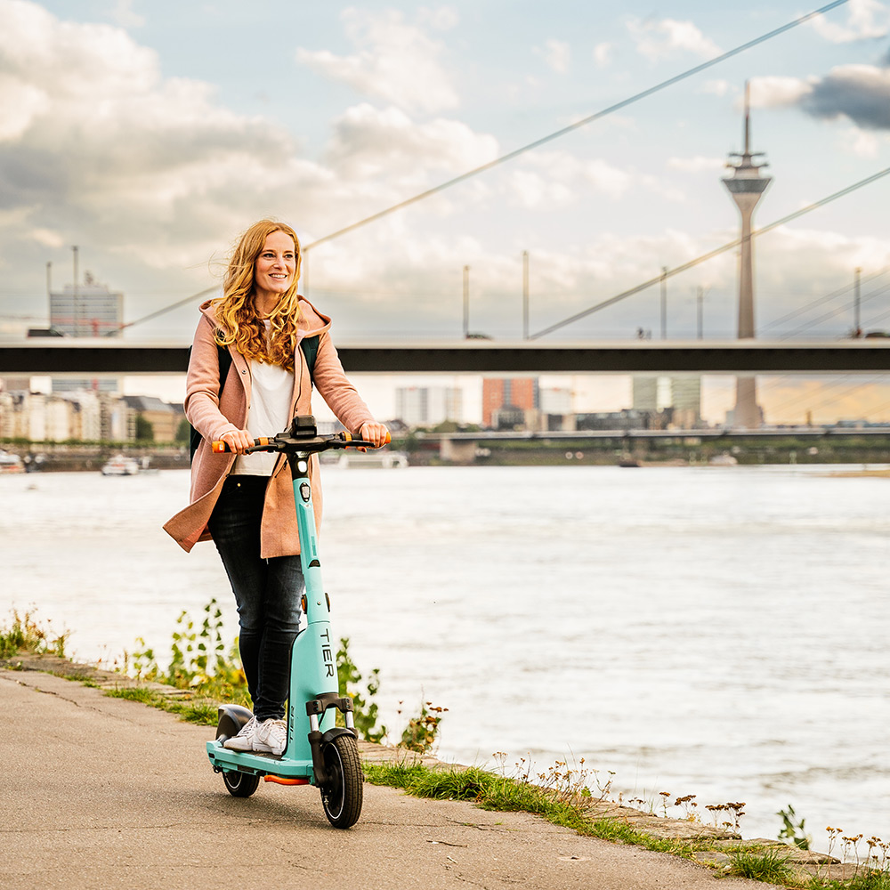 Rebecca fährt auf einem grünen E-Scooter am Rhein entlang, im Hintergrund ist der Düsseldorfer Fernsehturm zu sehen.