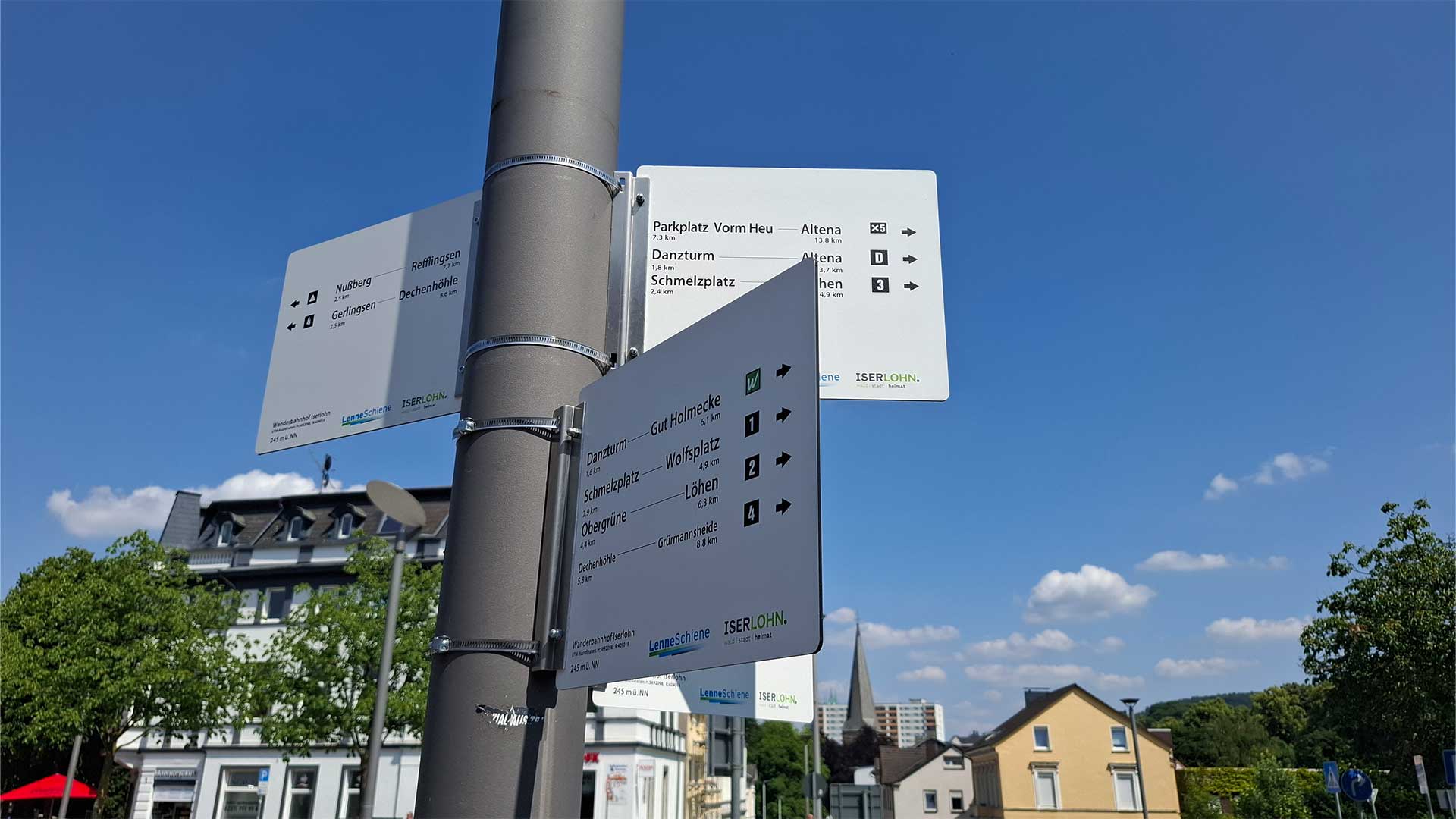 Wegweiser am Wanderbahnhof Iserlohn mit Zielen wie Danzturm, Dechenhöhle und Altena, blauer Himmel und Stadthäuser im Hintergrund