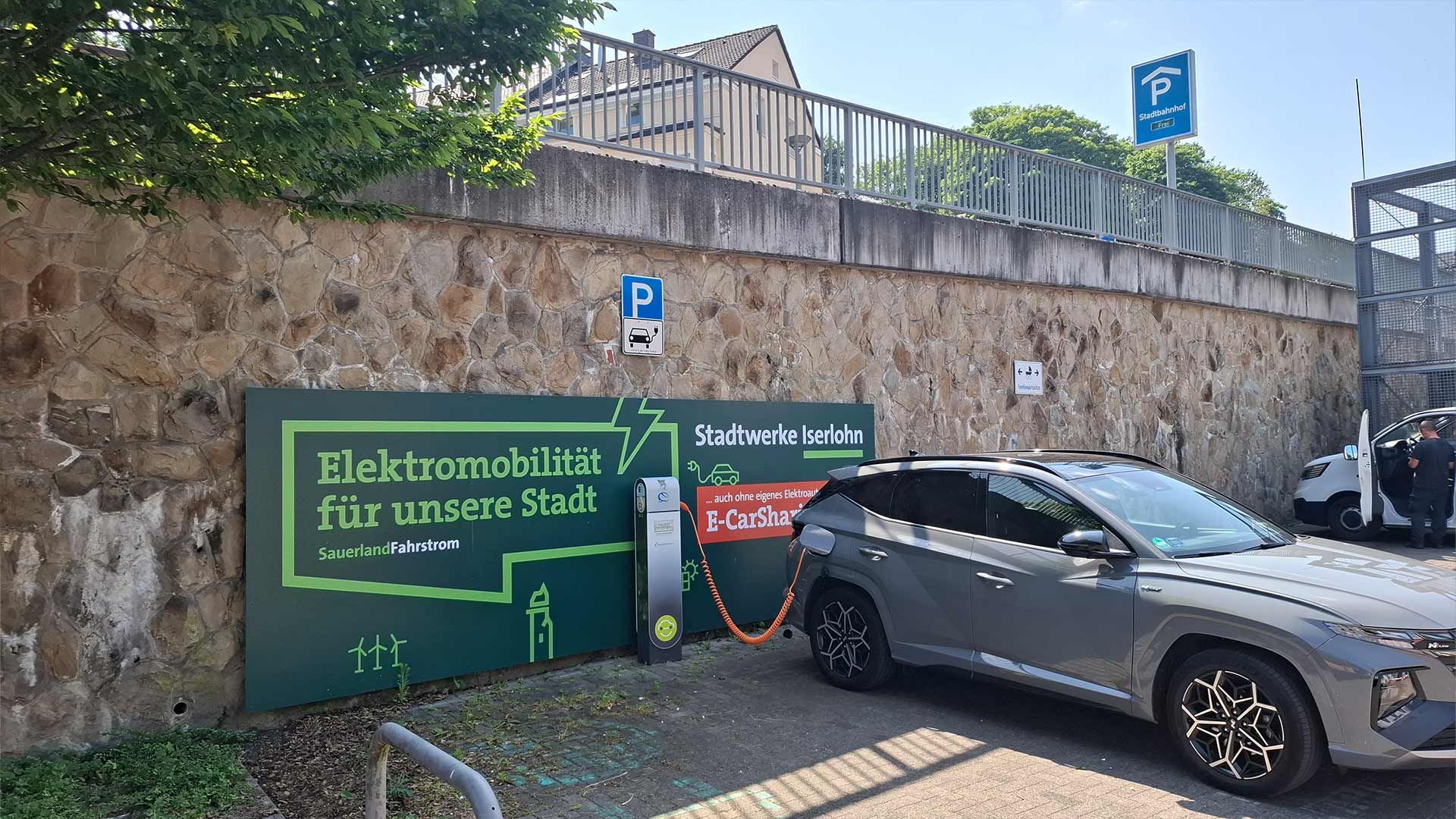 E-Auto lädt an öffentlicher Ladesäule der Stadtwerke Iserlohn am Bahnhof, Beschilderung „Elektromobilität für unsere Stadt“, sonniger Parkplatz mit Steinmauer