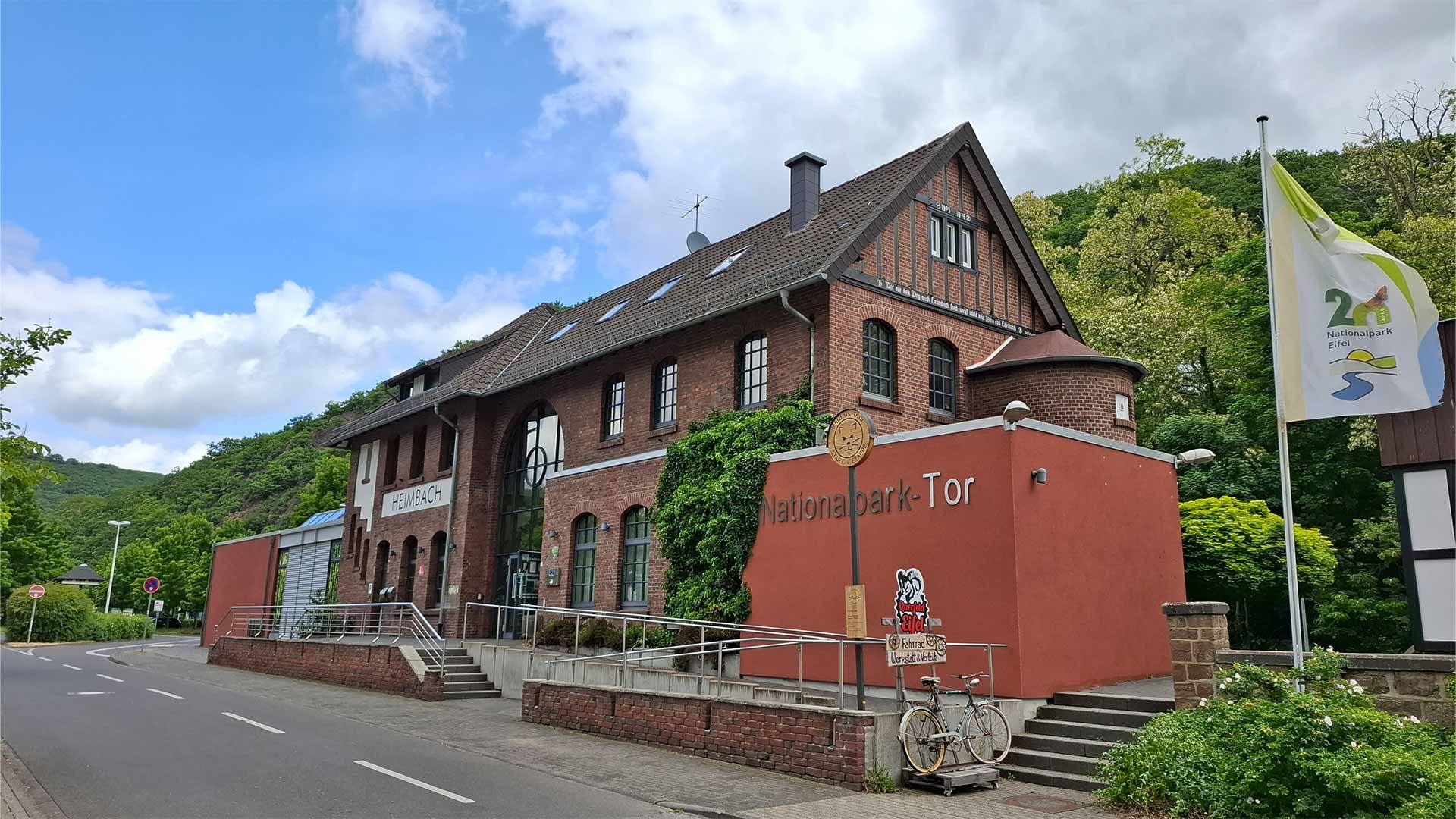 Nationalpark-Tor Heimbach im historischen Bahnhofsgebäude, rote Fassade mit Schriftzug, Fahrrad vor dem Eingang, grüne Eifel-Hügel im Hintergrund