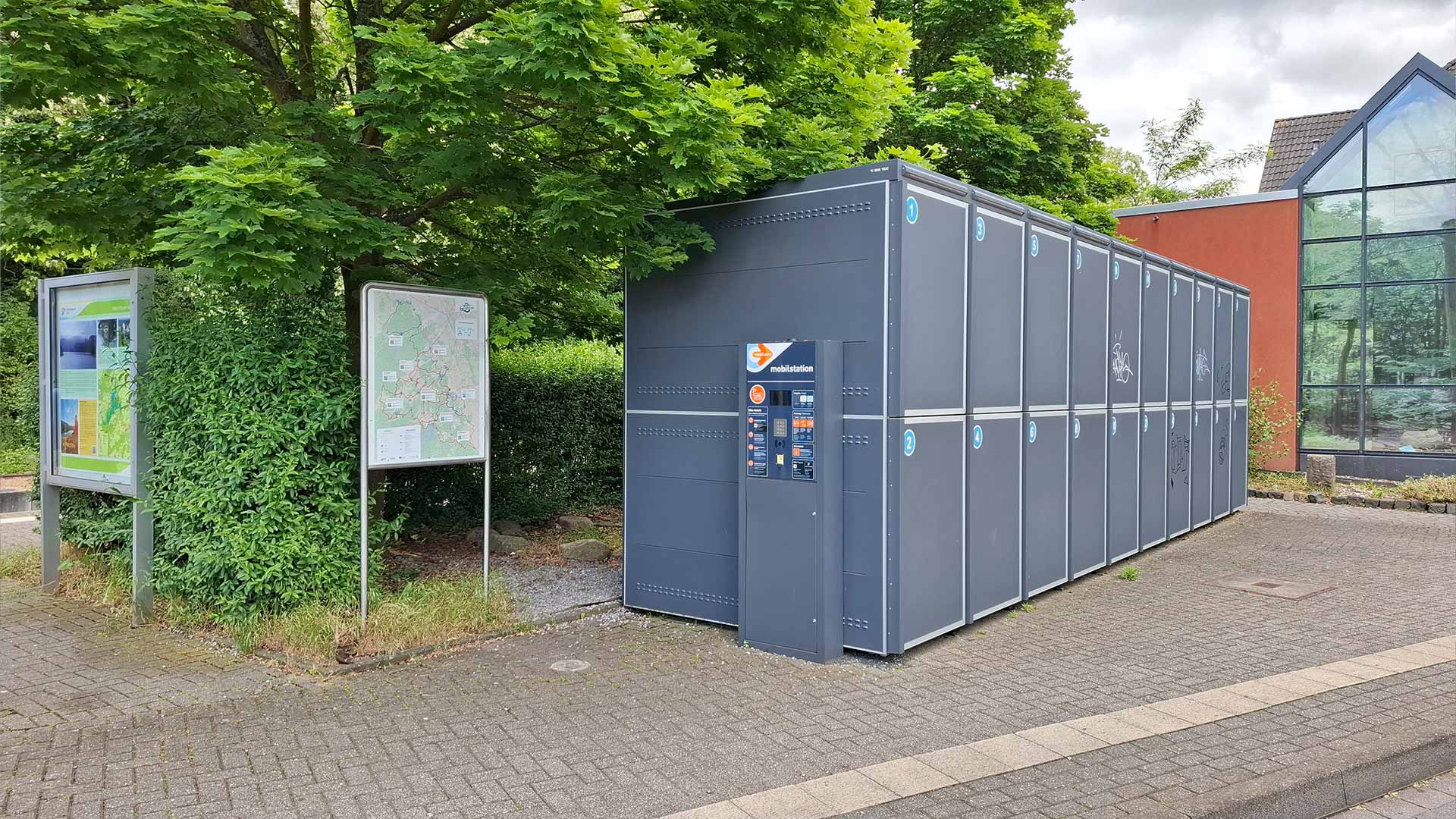 Moderne Fahrradboxen an der Mobilstation Heimbach neben Infotafeln und Stadtplan, graue Anlage unter Bäumen, sichere Abstellmöglichkeiten für Radreisende