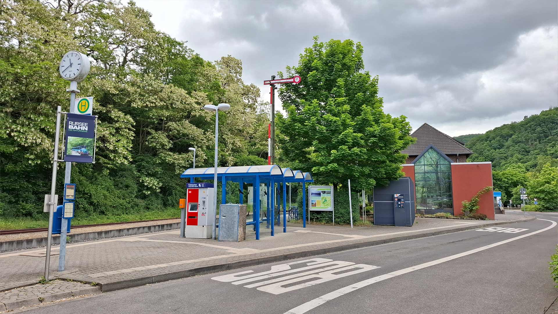 Haltepunkt Heimbach mit blauem Wartehäuschen, Ticketautomat und Bushaltestelle, umgeben von viel Grün und Hinweistafeln, ruhige Kleinstadtatmosphäre