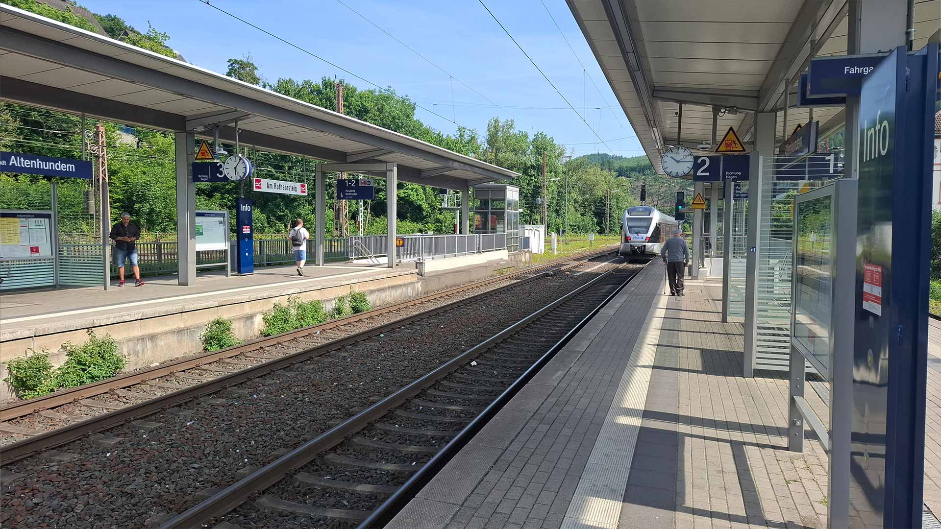 Moderner Bahnhof Lennestadt‑Altenhundem mit einfahrendem Regionalzug, überdachte Bahnsteige, Fahrgäste warten, grüne Hügellandschaft im Hintergrund