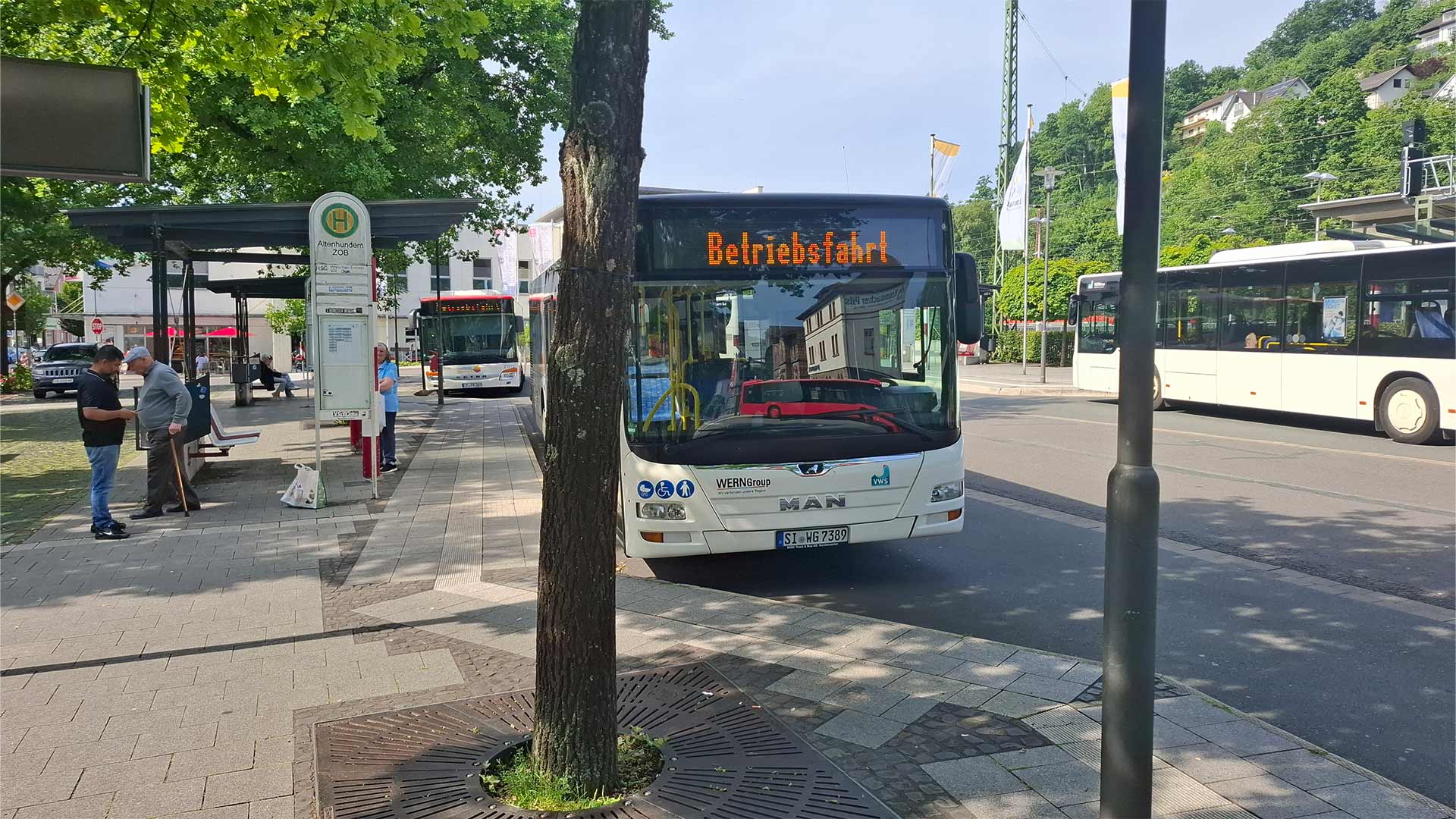 Busbahnhof Lennestadt‑Altenhundem mit mehreren Linienbussen und Fahrgästen an der Haltestelle, grüner Baumbestand und Stadtbebauung