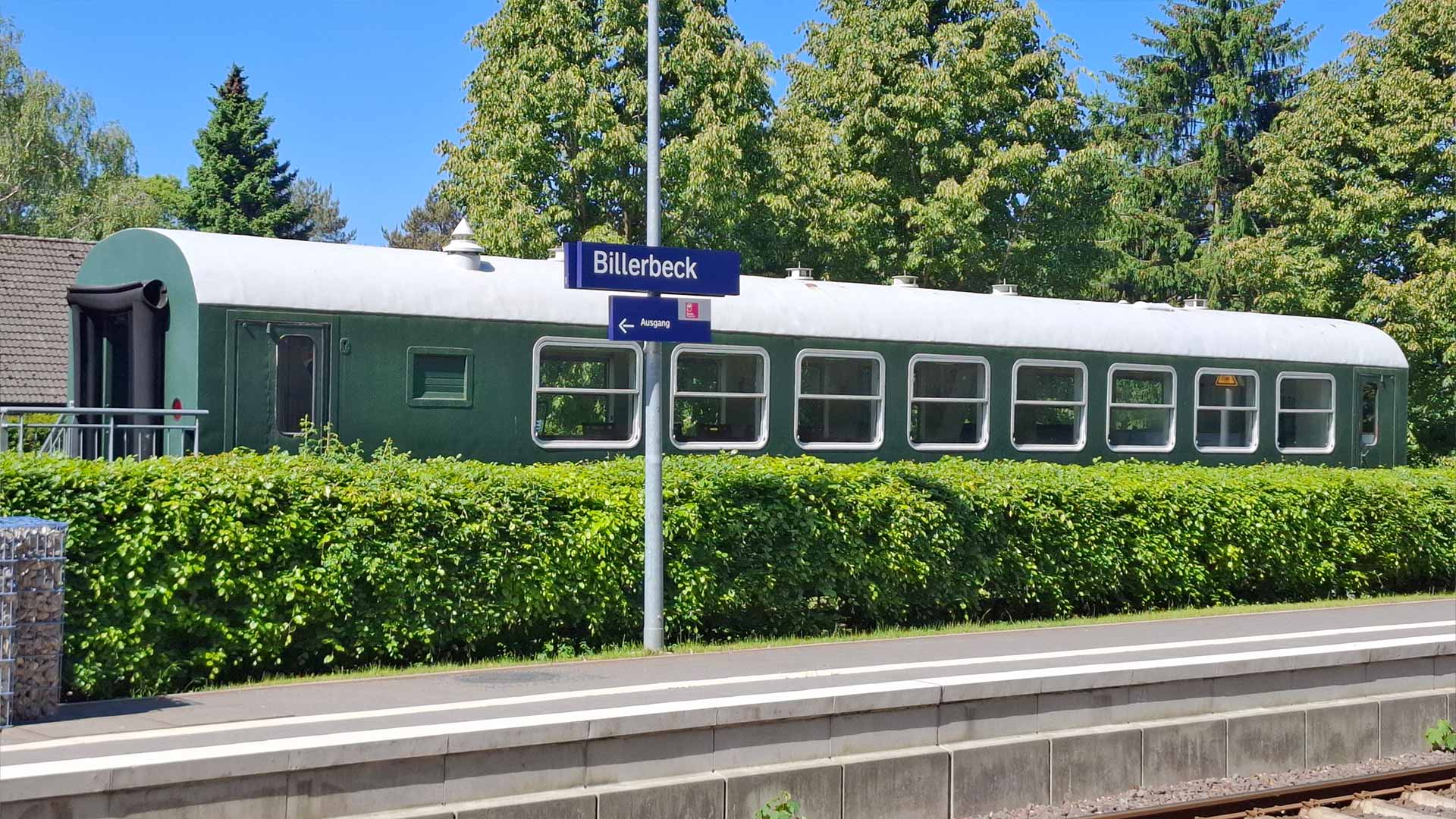 Grüner historischer Bahnwagen am Bahnhof Billerbeck hinter Hecke, Stationsschild „Billerbeck“, sommerliche Bäume im Hintergrund