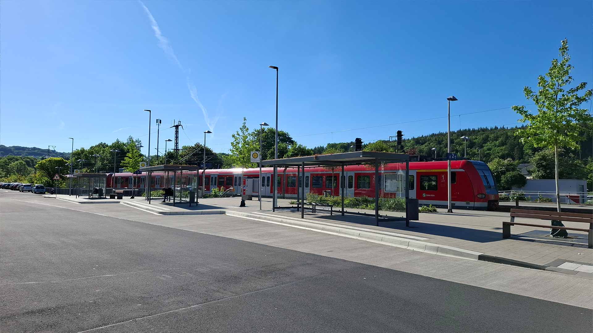 Bus‑ und Umstiegsbereich am Bahnhof Schladern mit Unterständen, dahinter roter S‑Bahn‑Zug und grüne Hügel