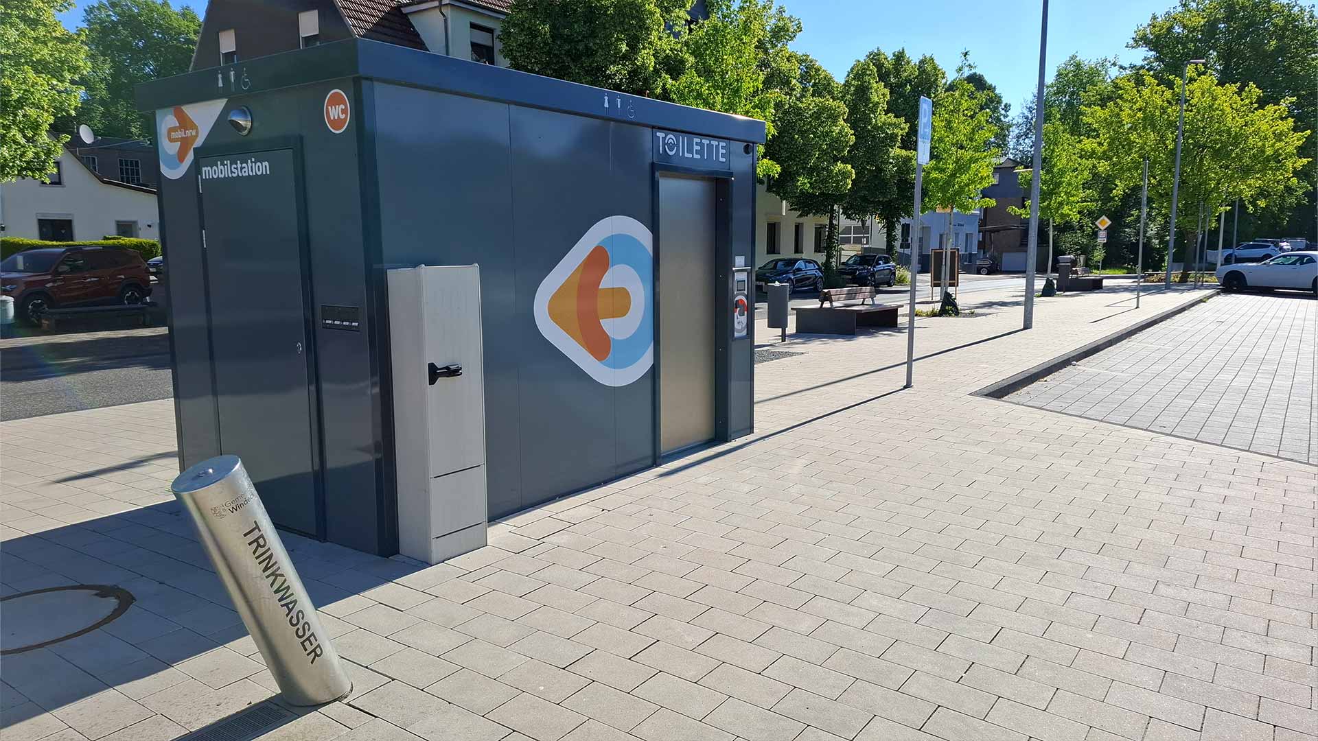 Öffentliche WC‑Anlage an der Mobilstation Schladern mit Trinkwasser‑Zapfstelle, moderner Platz mit Sitzbänken und Bäumen