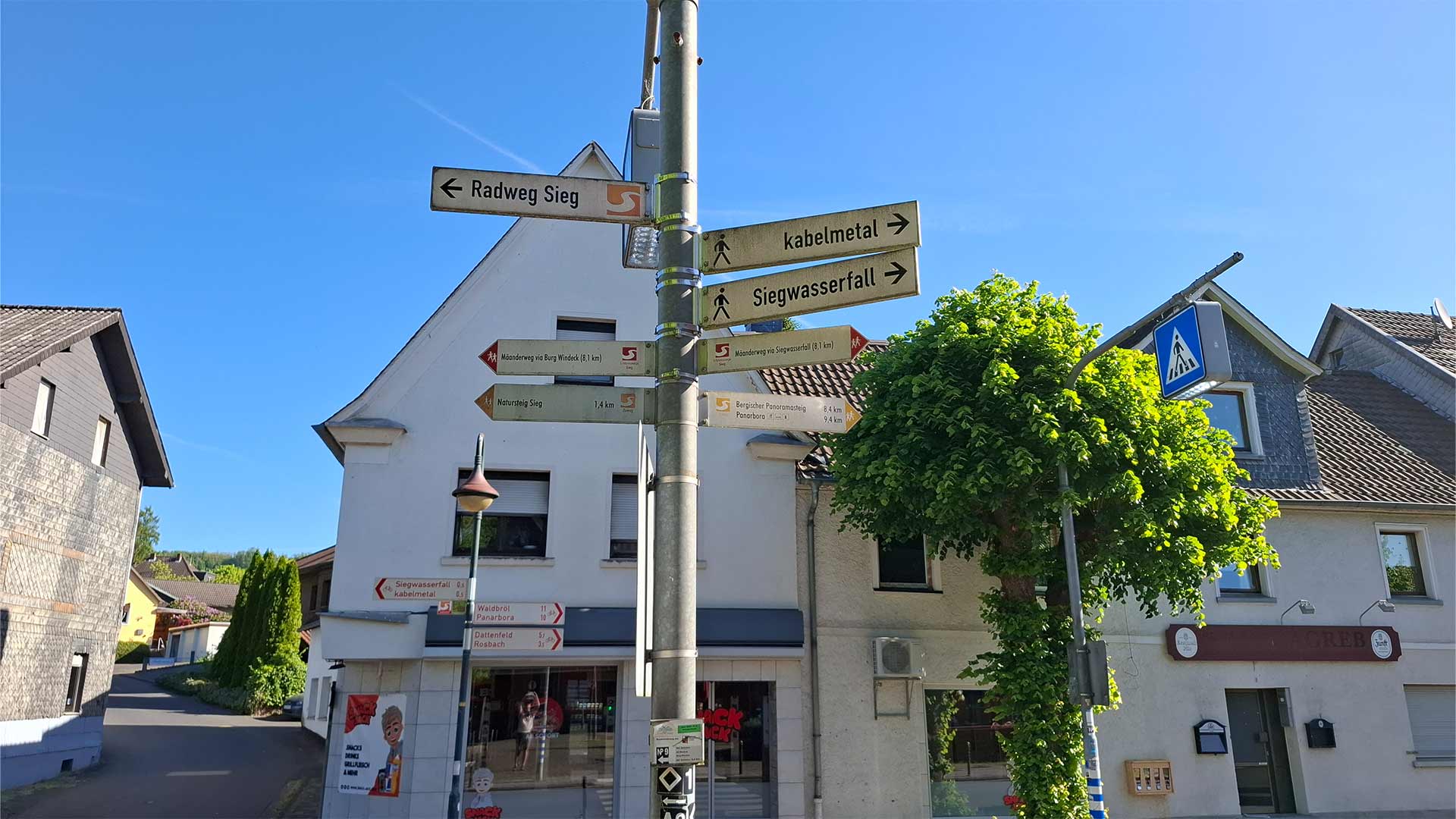 Wegweiser im Ortskern Schladern zu Siegfall, kabelmetal und Radweg Sieg, blau­er Himmel über Fachwerk‑ und Wohnhäusern