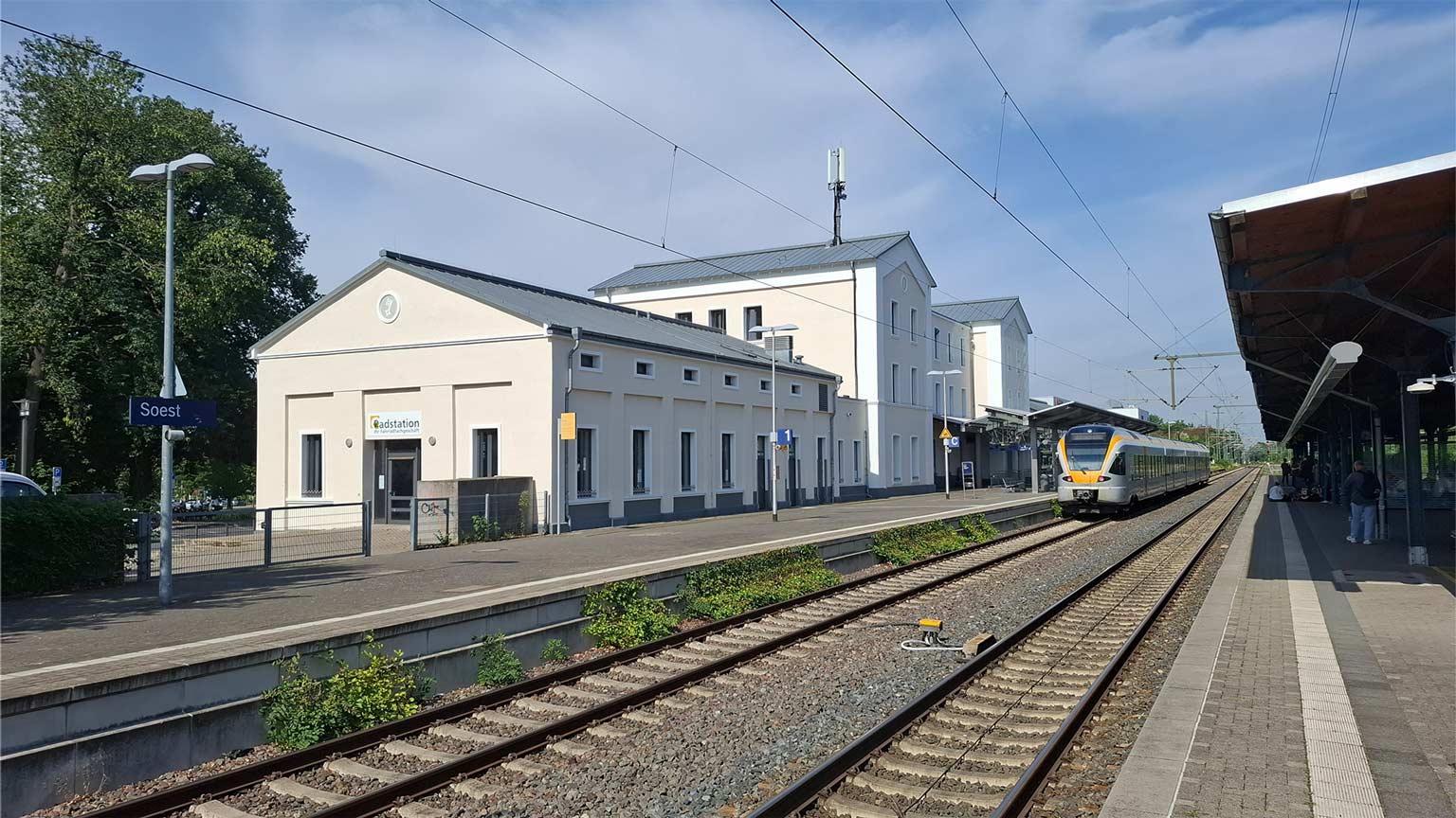 Heller Bahnhof Soest mit Regionalzug am Gleis, historische Gebäude entlang der Bahnsteige, Oberleitungen und weiter Blick über die Gleise