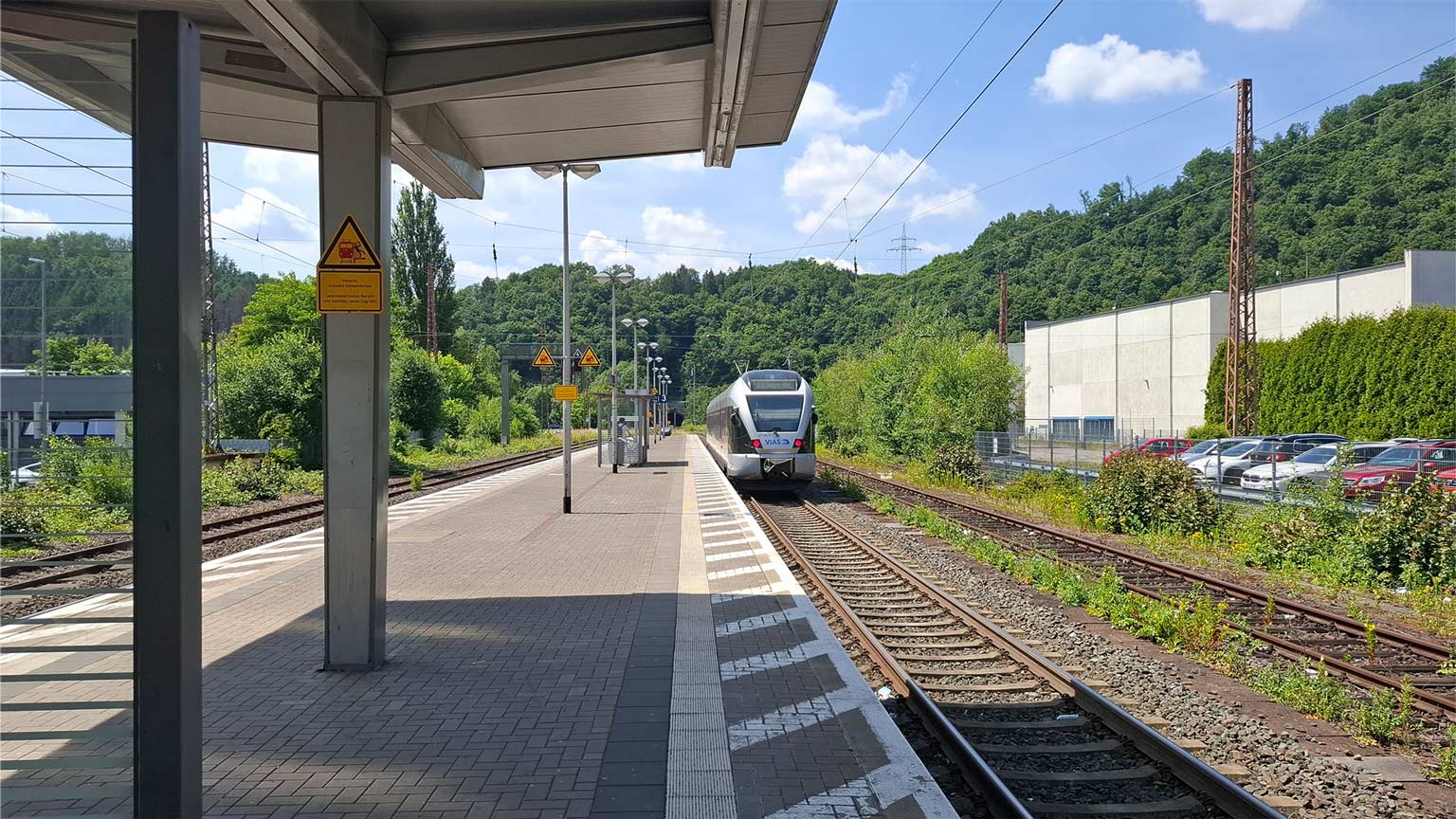 Regionalzug am Bahnhof Werdohl fährt ein, überdachte Bahnsteige, grüne Hänge und Industrieareal am Rand, Sommerhimmel