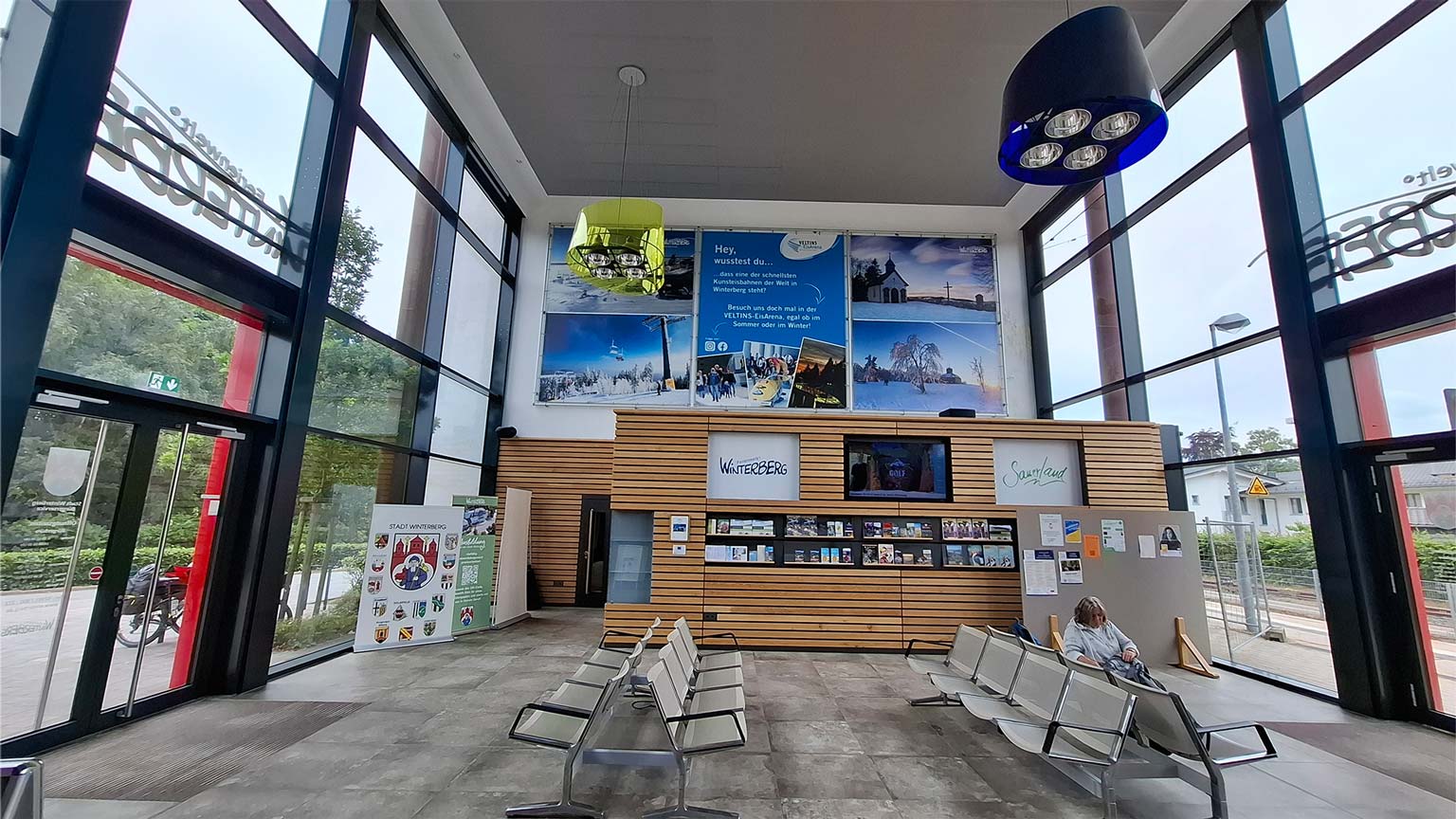 Helle Wartehalle im Bürgerbahnhof Winterberg mit Sitzreihen, Tourist‑Information und großen Panoramafenstern, Infowände zu Winter‑ und Sommeraktivitäten