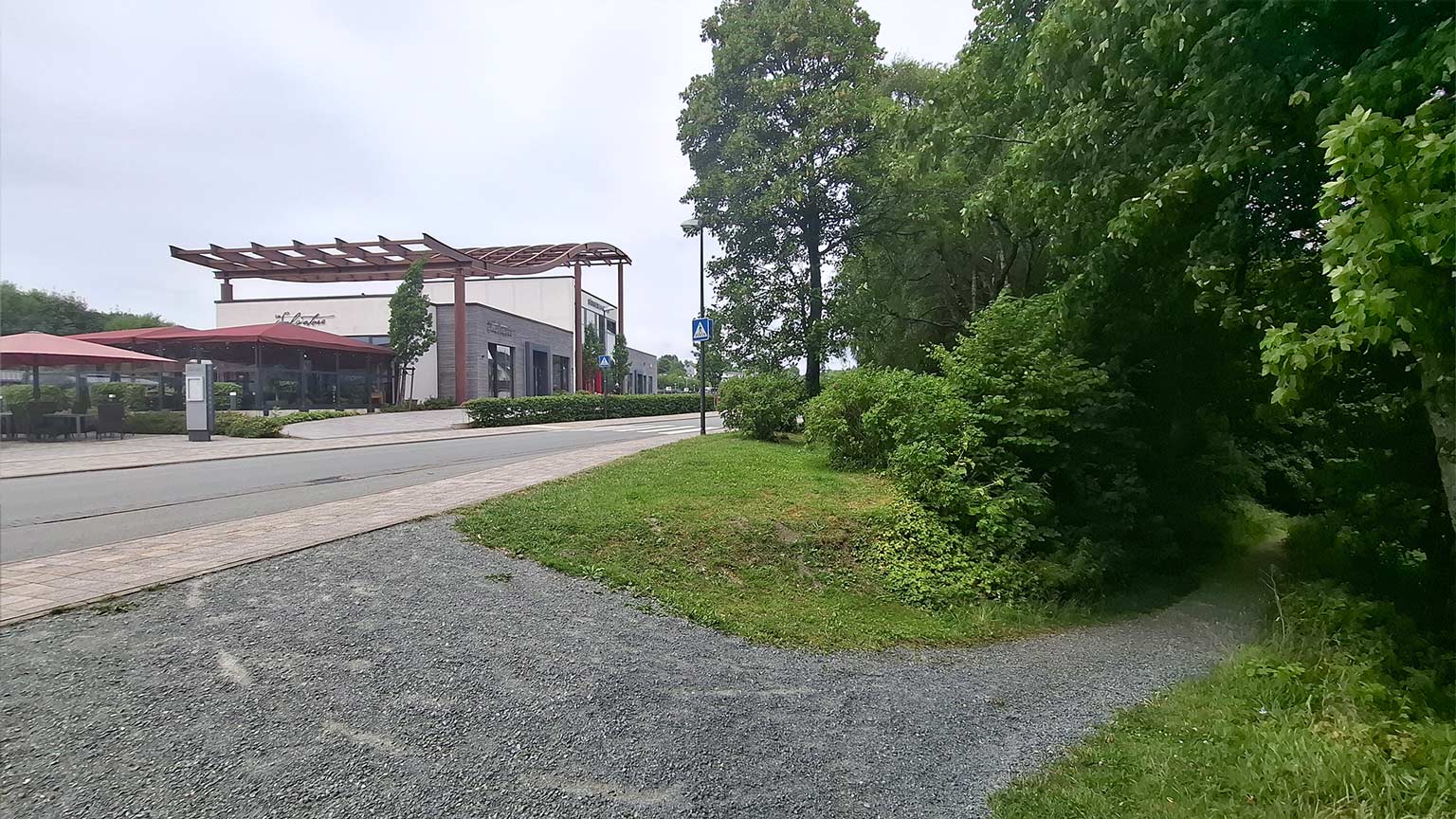 Übergang vom Bürgerbahnhof Winterberg zum Wanderweg; modernes Gebäude mit Terrasse links, rechts beginnt ein schmaler Pfad in den grünen Wald