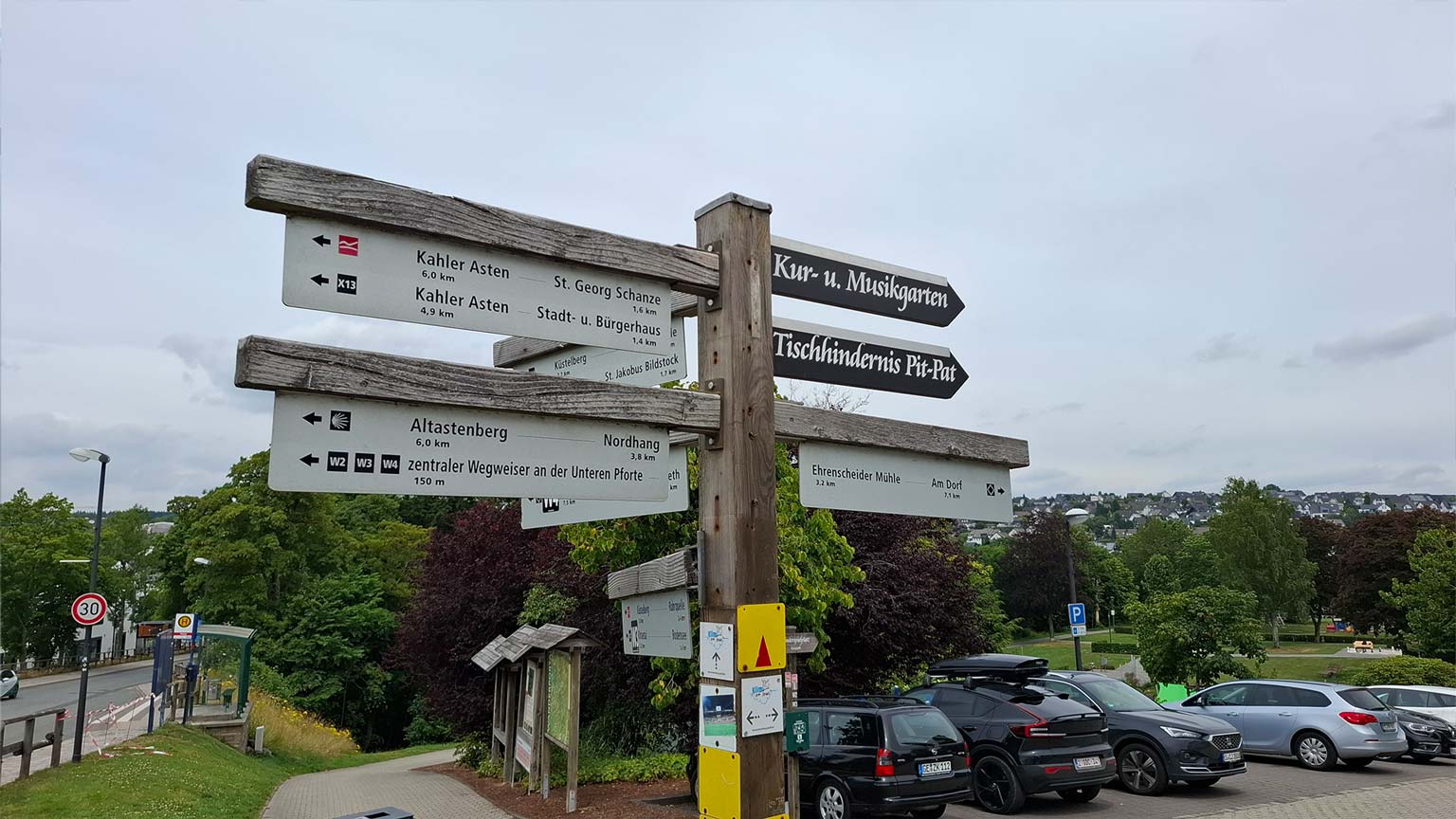 Wegweiser am Bürgerbahnhof Winterberg zu Zielen wie Kahler Asten, Altastenberg und Kur‑ und Musikgarten, Blick über Parkplätze und Grünflächen