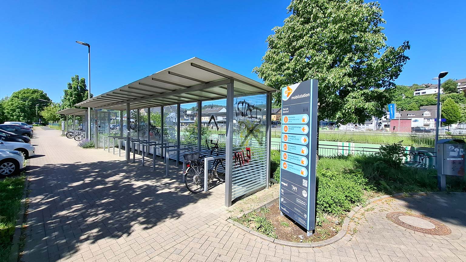 Überdachte Fahrradabstellanlage an der Mobilstation Overath, Infostele mit Angeboten wie Carsharing und ÖPNV, Parkplätze und Grünflächen