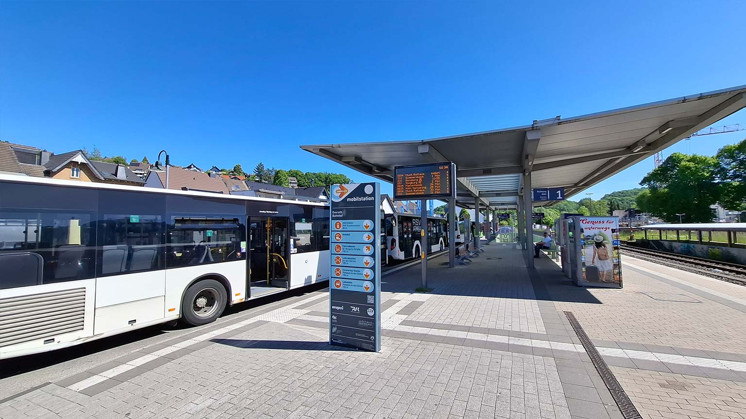 Busbahnhof Overath mit mehreren Linienbussen, digitaler Abfahrtsanzeige und Mobilstations‑Hinweistafel, direkte Umsteigemöglichkeit zu den Bahnsteigen