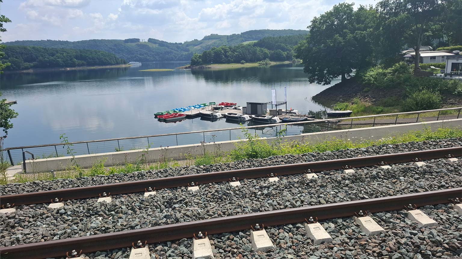 Blick vom Bahnhof Sondern auf den Biggesee mit Bootsverleih und Stegen, ruhiges Wasser und bewaldete Uferlandschaft