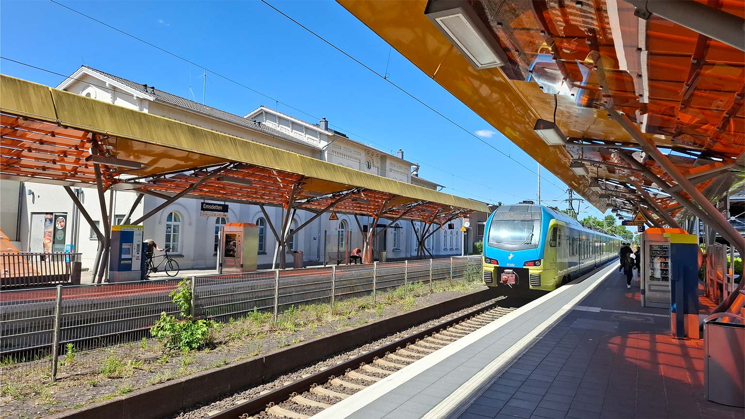 Bahnsteig Emsdetten mit einfahrendem Regionalzug, orange‑gelbe Dächer, Ticketautomaten und Reisenden bei Sonnenschein