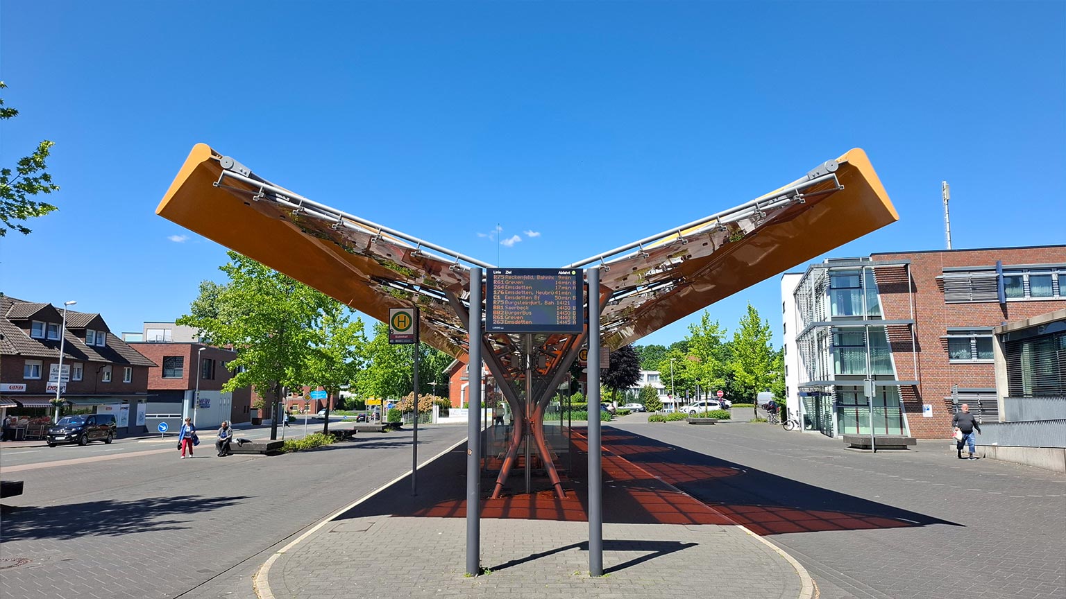 Zentraler Bussteig am Bahnhof Emsdetten mit markanter V‑förmiger Überdachung und digitaler Abfahrtsanzeige, Stadtquartier im Hintergrund