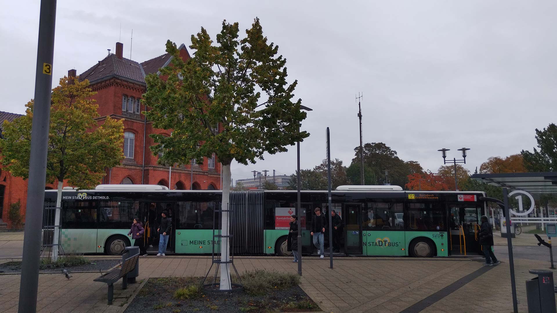 Busbahnhof am Bahnhof Detmold mit Gelenkbus „Meine Stadtbus Detmold“, herbstliche Bäume und Backsteinfassade im Hintergrund