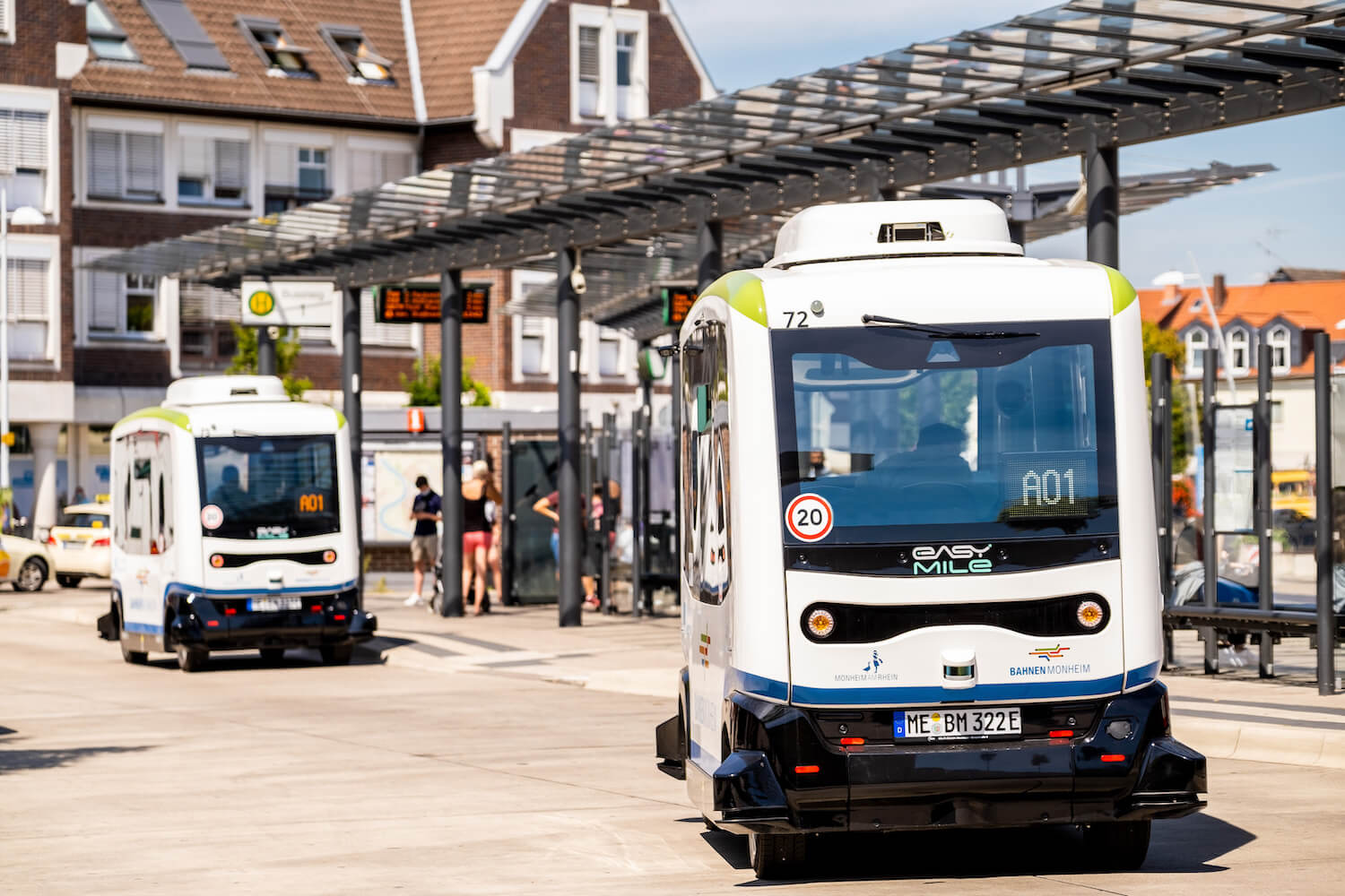 Zwei hintereinander fahrende autonome Kleinbusse an einem Busbahnhof.