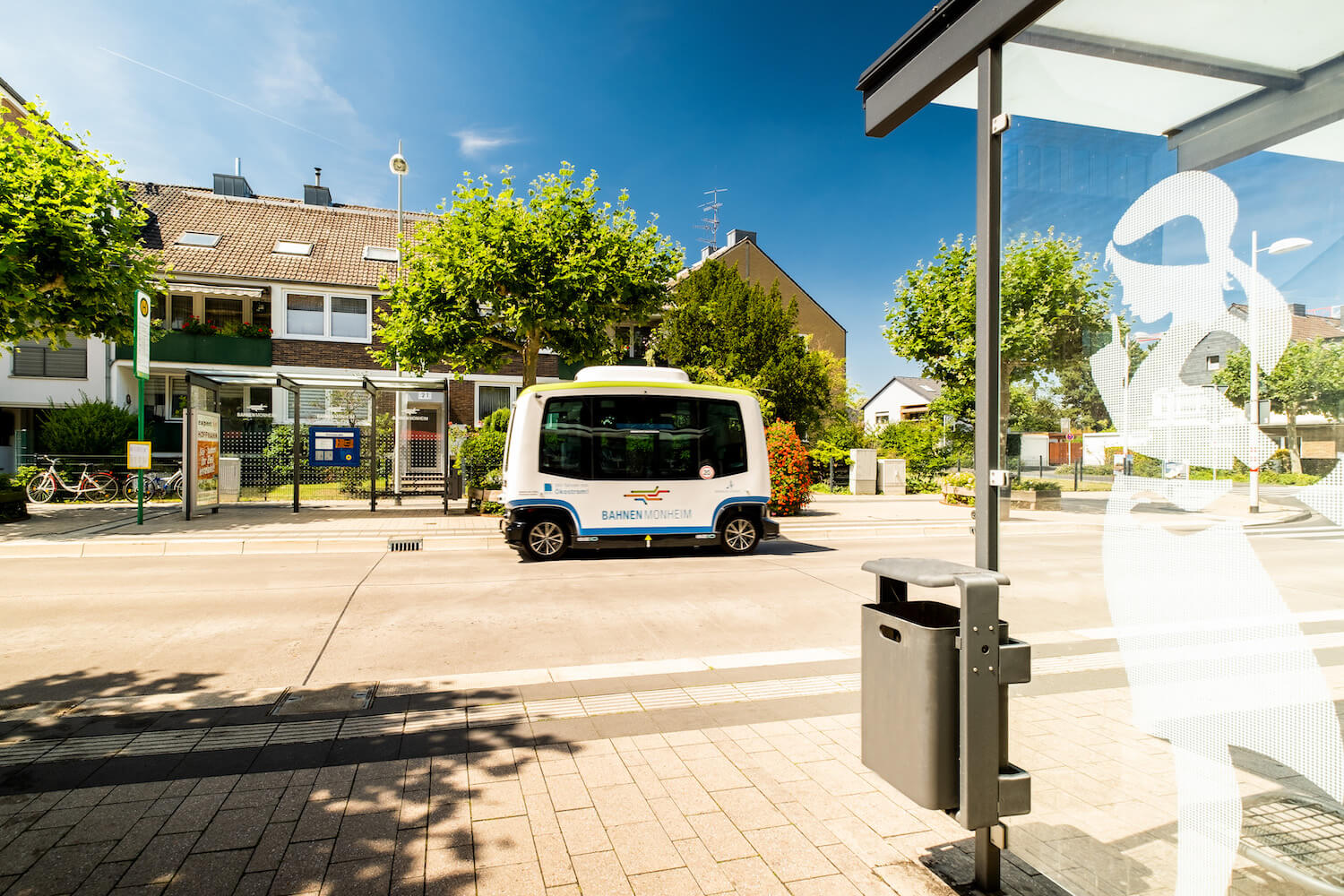 Ein autonomer Kleinbus fährt an einem sonnigen Tag eine Straße entlang, an der Bushaltestellen zu sehen sind.