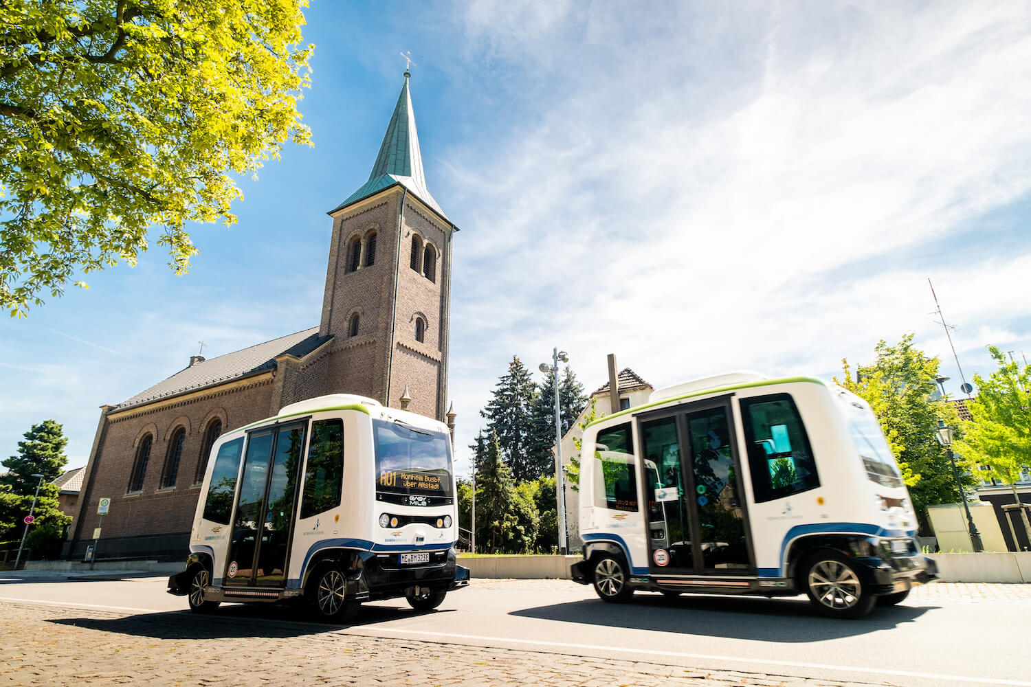 Zwei autonome Kleinbusse fahren auf Kopfsteinpflaster an einem sonnigen Tag, im Hintergrund ist eine Kirche zu sehen.