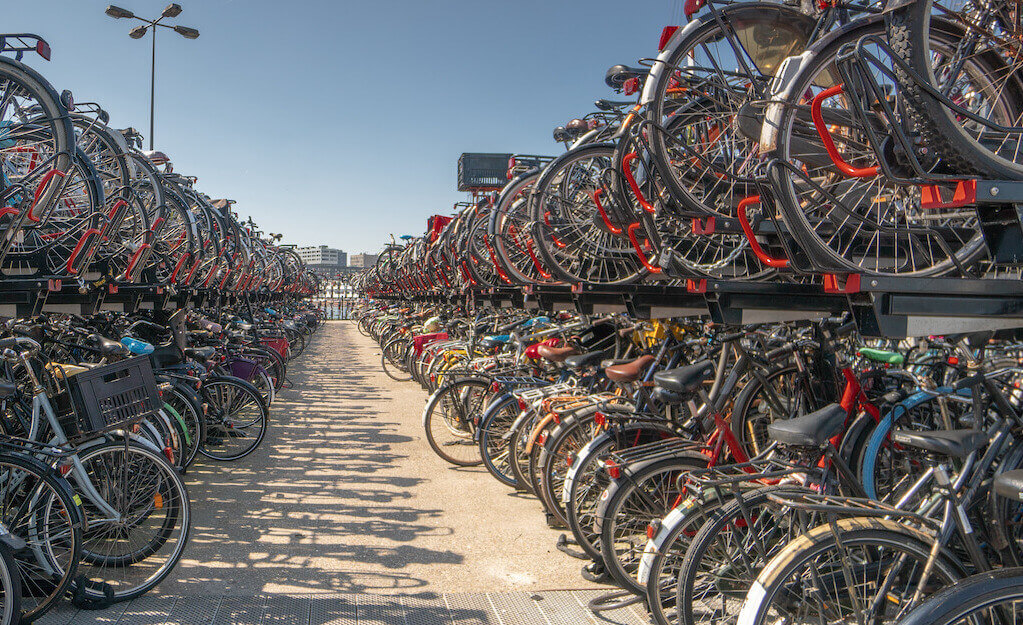 Seitlicher Blick bei blauem Himmel auf die Fahrrad-Stellplätze im Zentrum Amsterdams. Links und rechts hängen und stehen Massen an Fahrrädern an Halterungen, es ist kaum freier Platz zwischen den Rädern.