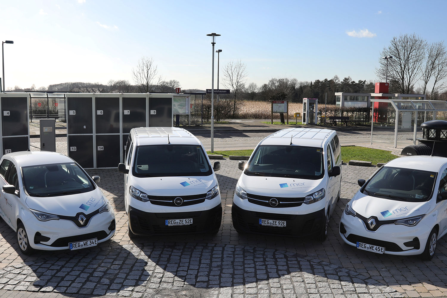 Vier E-Autos stehen auf einem Kopfsteinplatz vor den Gleisen des Bahnhofs Borgholzhausen.