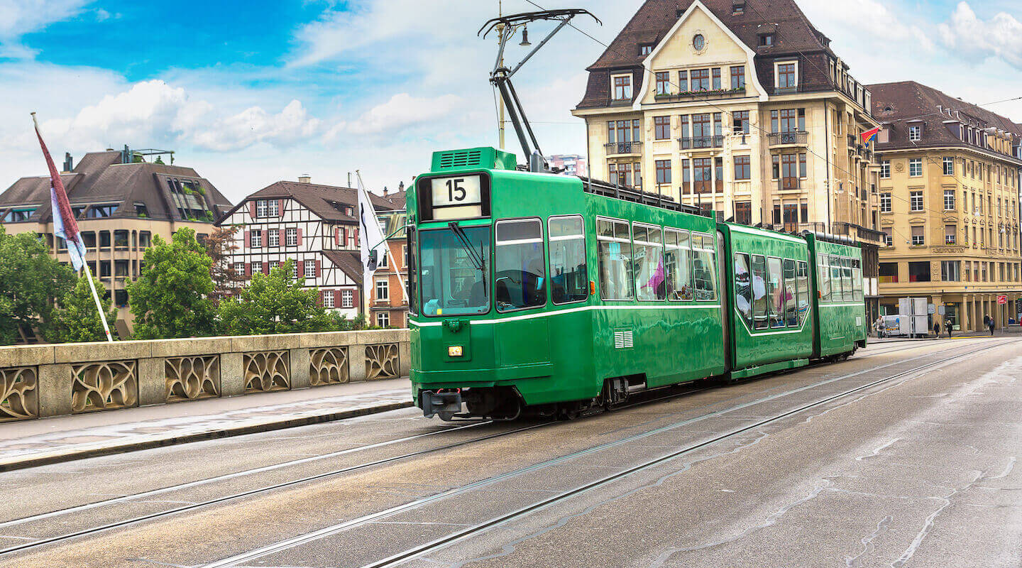 Eine grüne Straßenbahn fährt auf Schienen. Im Hintergrund sieht man Fachwerkhäuser und andere Bauten, die zu Basel gehören.