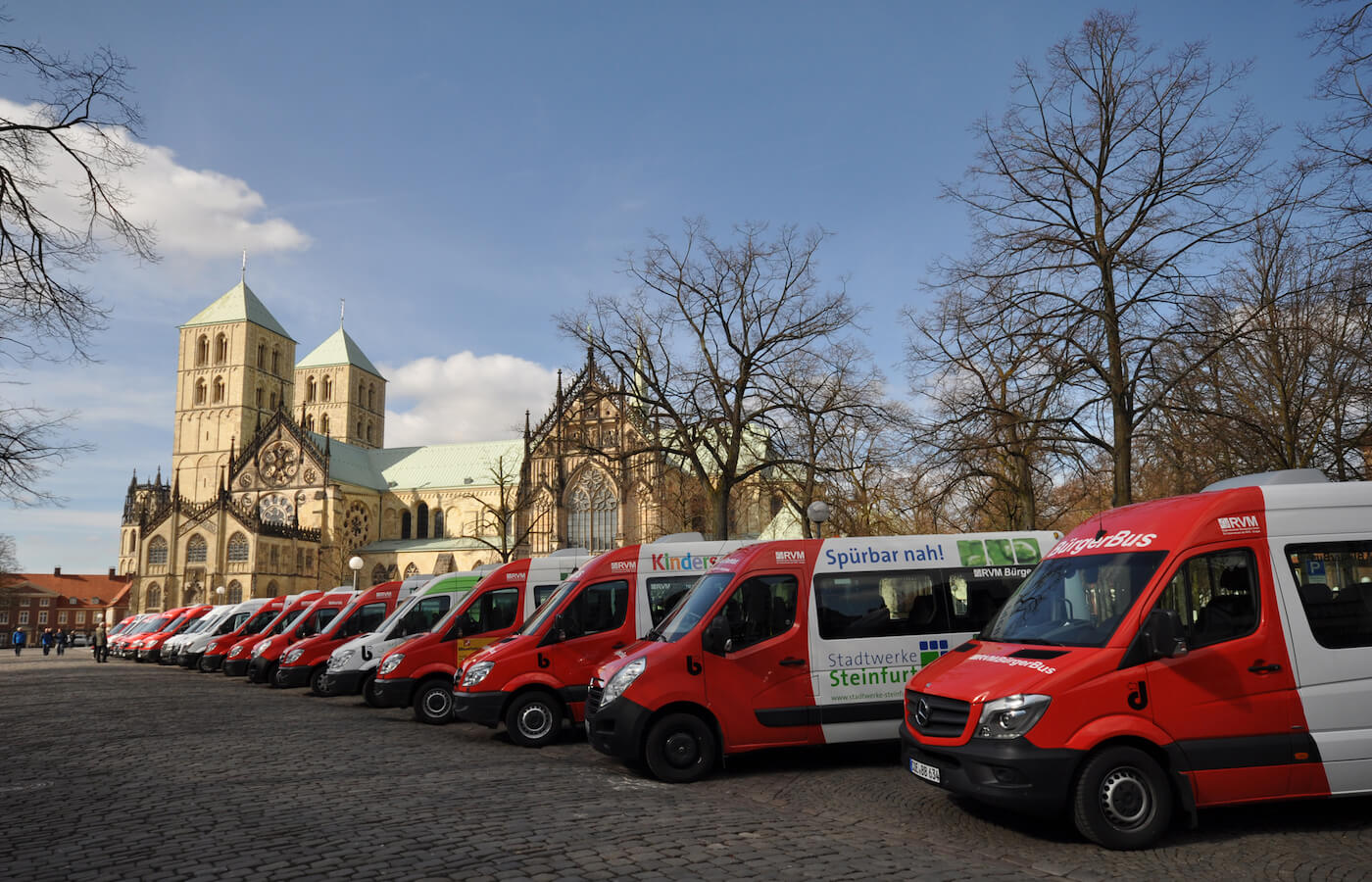 Eine Reihe roter Kleinbusse steht nebeneinander auf einem Parkplatz, im Hintergrund sieht man ein altes Gebäude.