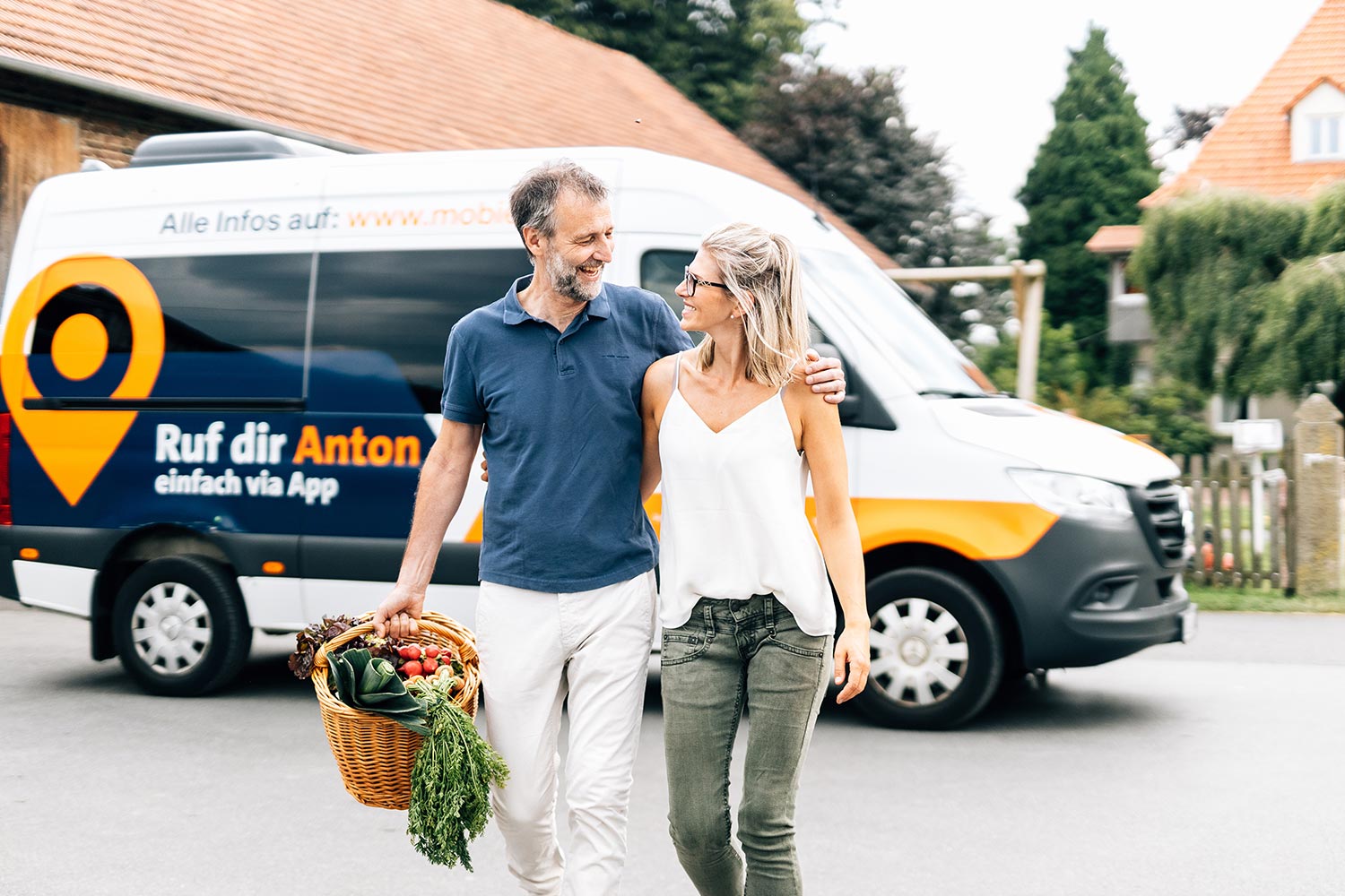 Ein Ehepaar mittleren Alters steht mit einem Einkaufskorb voller Gemüse vor dem Rufbus Anton.