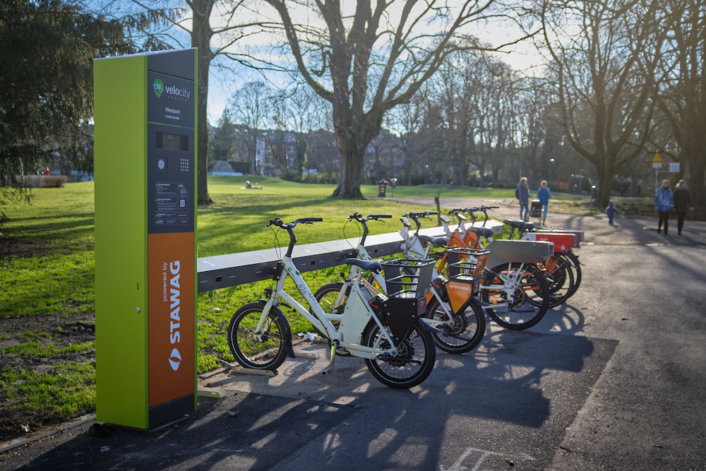 E-Bikes stehen in einer Reihe an einer Verleihstation vor einer Wiese.