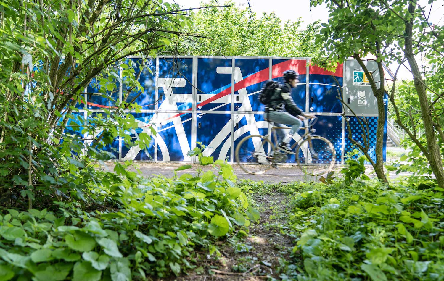 Ein Mann fährt auf einem Fahrrad an einer blauen Radbox vorbei.