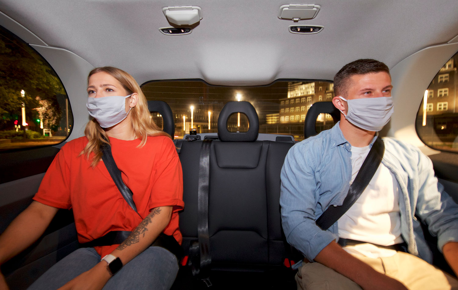 Eine junge Frau und ein junge Mann sitzen mit Mund-Nasen-Bedeckung nebeneinander in einem Auto.