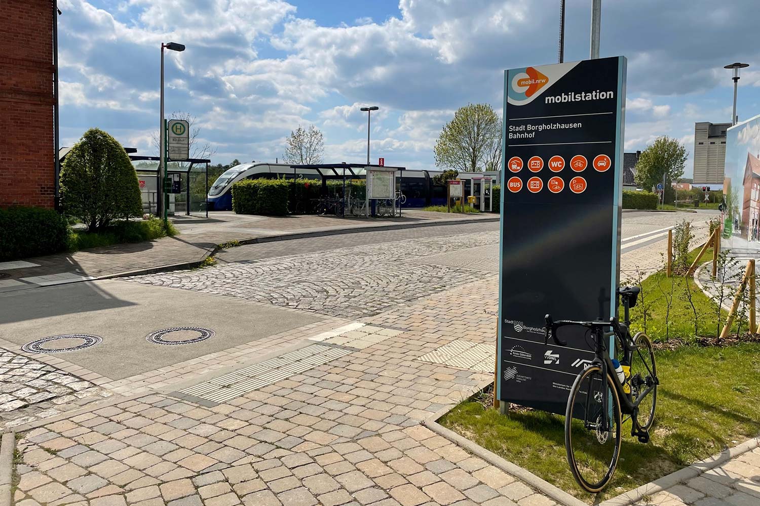 Ein Fahrrad lehnt an der Stele der Mobilstation Borgholzhausen, während im Hintergrund ein Zug fährt.