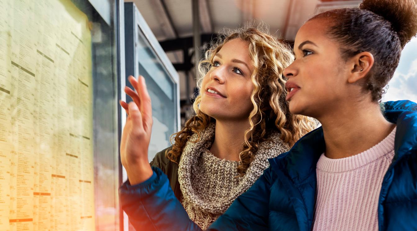Header Fahrplanauskunft Zwei junge Frauen stehen an einem Bahnsteig und betrachten den Abfahrtsplan.