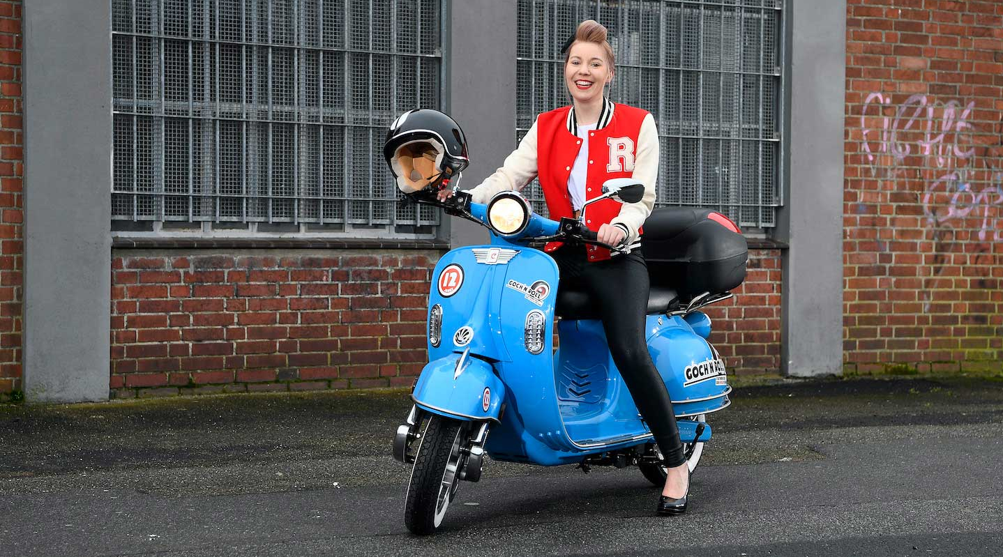 Eine junge Frau im Rockabilly-Style sitzt auf einem blauen E-Roller.