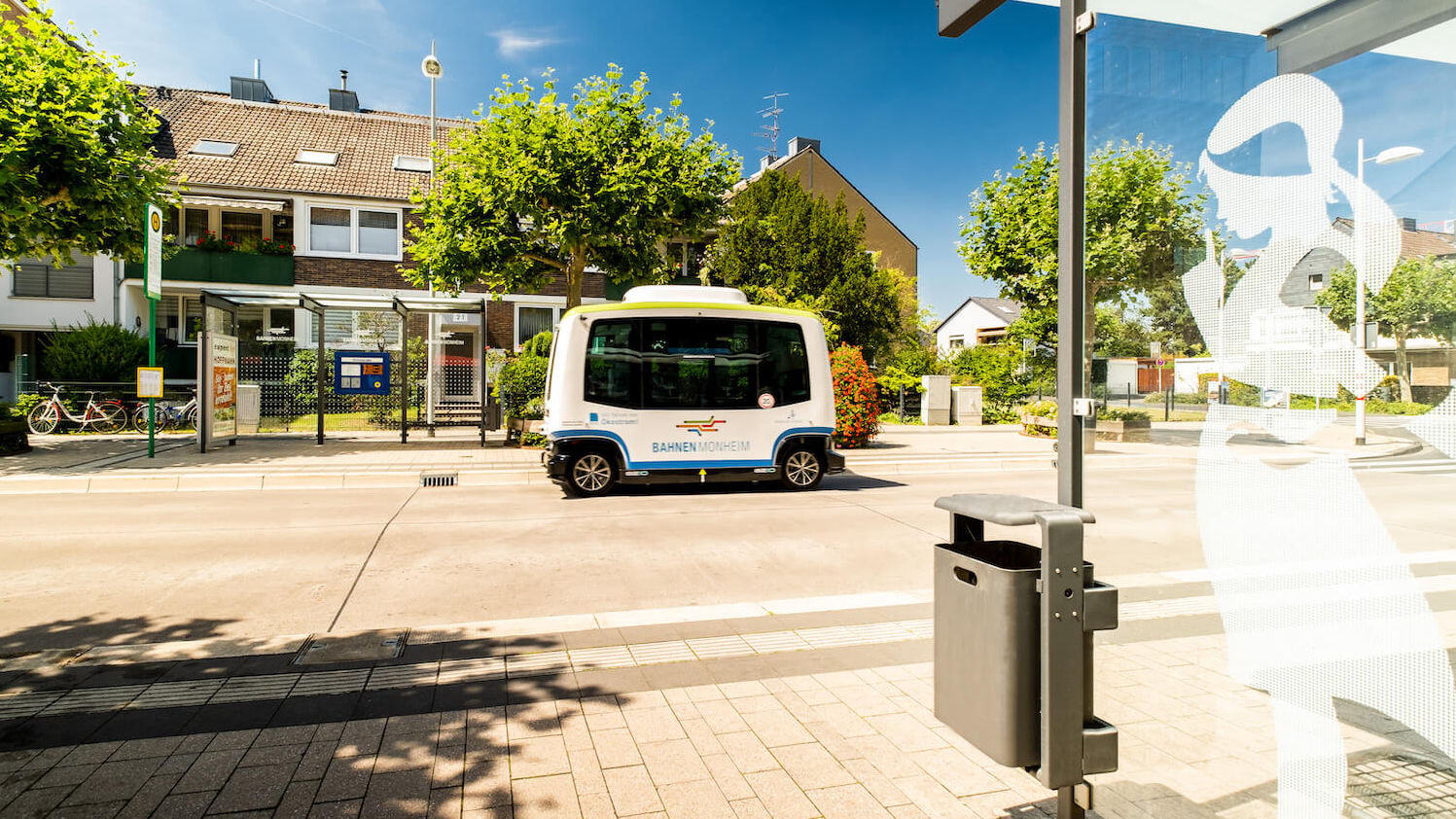 Ein autonomer Kleinbus fährt an einem sonnigen Tag eine Straße entlang, an der Bushaltestellen zu sehen sind.
