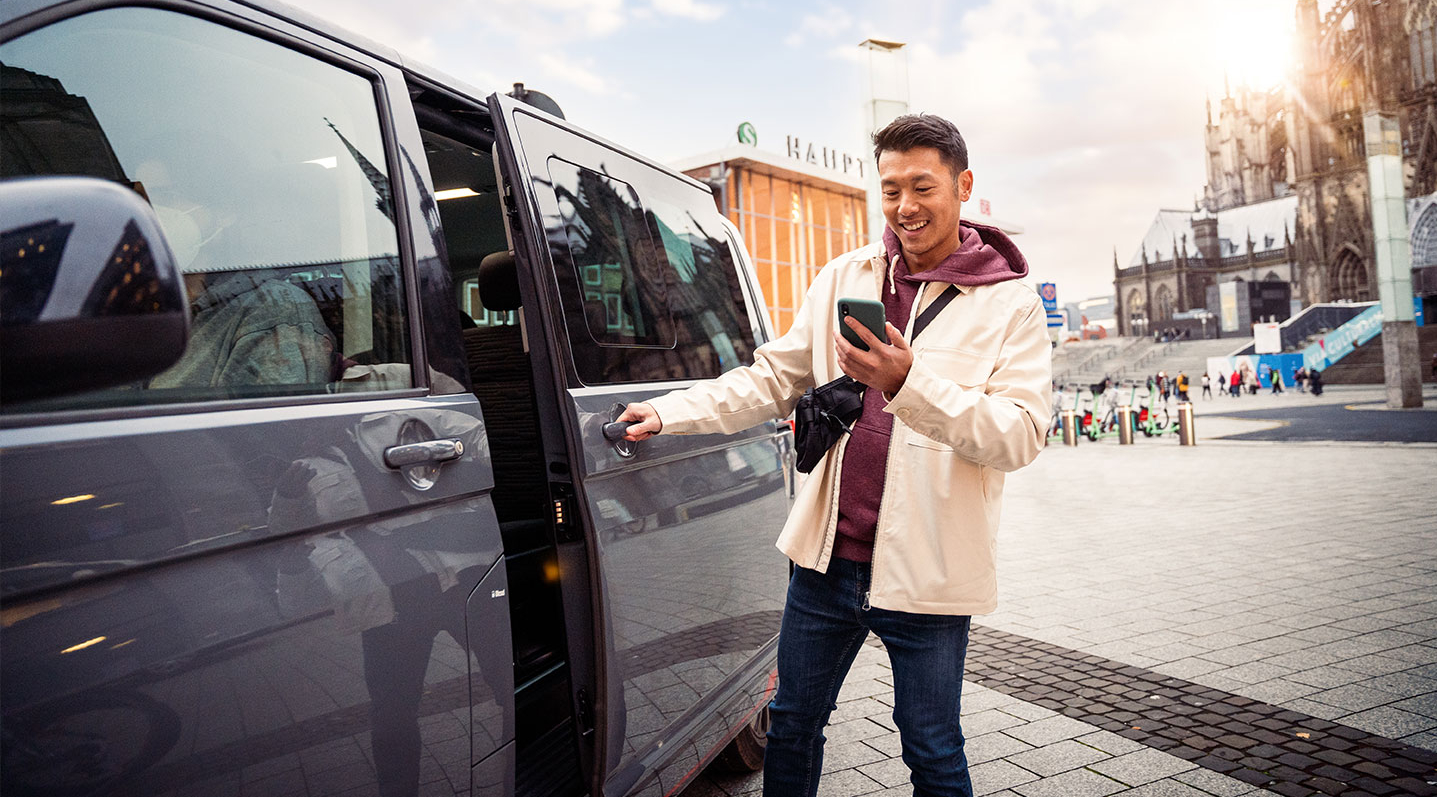 Ein junger Mann steigt in ein On-Demand-Fahrzeug und schaut dabei auf sein Handy.