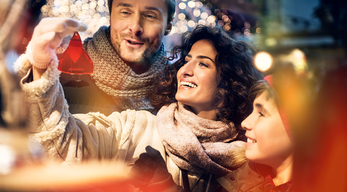 Eine Familie mit Mutter, Vater und Sohn steht an einem Weihnachtsmarktstand und schaut sich fröhlich die Waren an.
