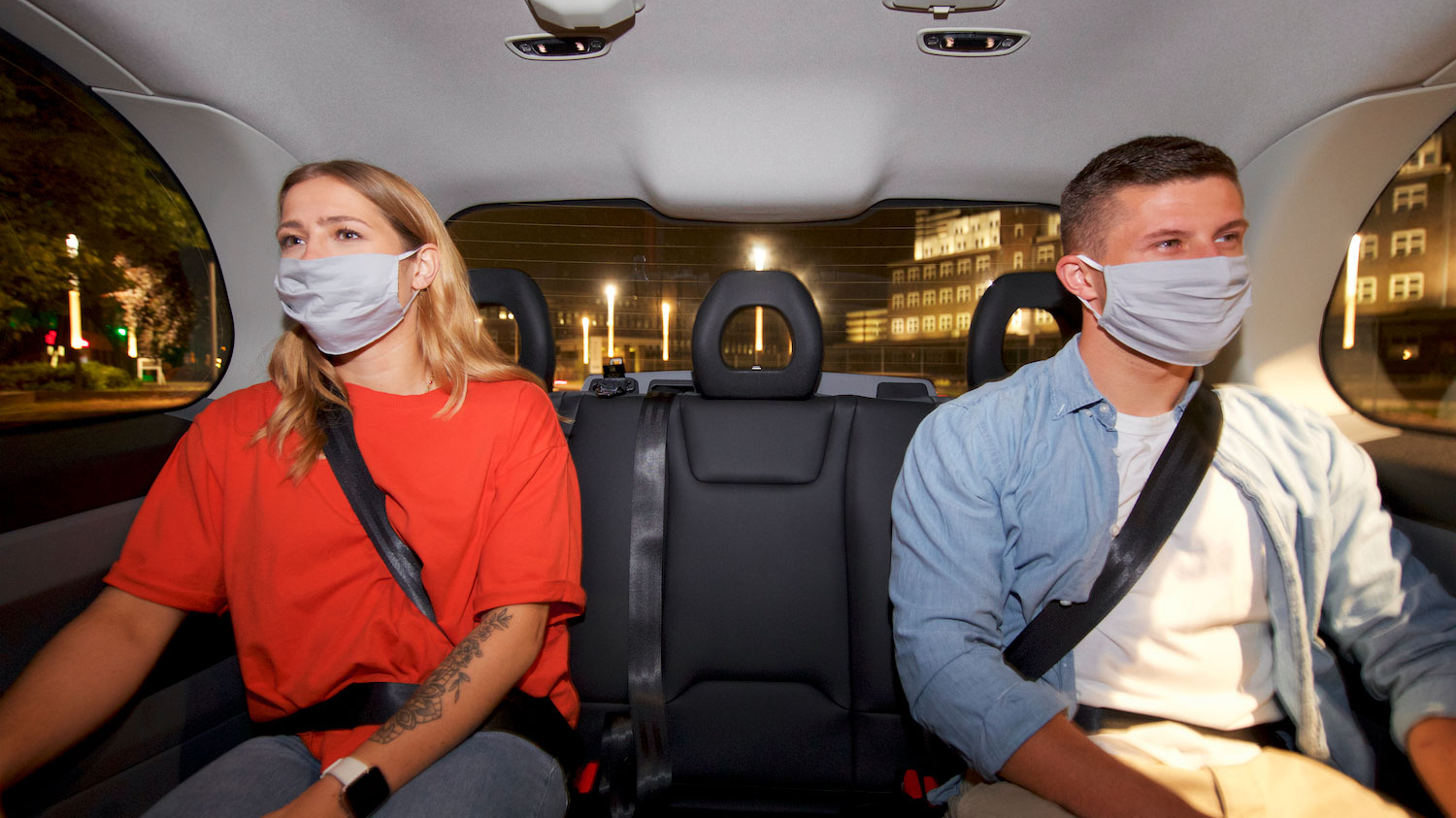 Eine junge Frau und ein junge Mann sitzen mit Mund-Nasen-Bedeckung nebeneinander in einem Auto.
