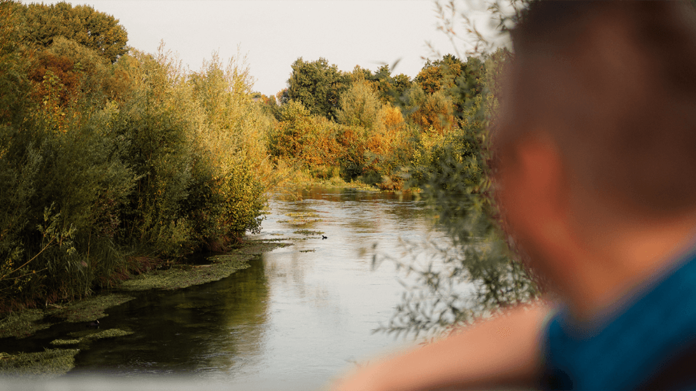 Zu sehen ist ein Landschaftbild mit Fluss und Wald. Im Vordergrund erkennt man die Silhouette eines Oberkörpers eines Mannes in unschärfe. 