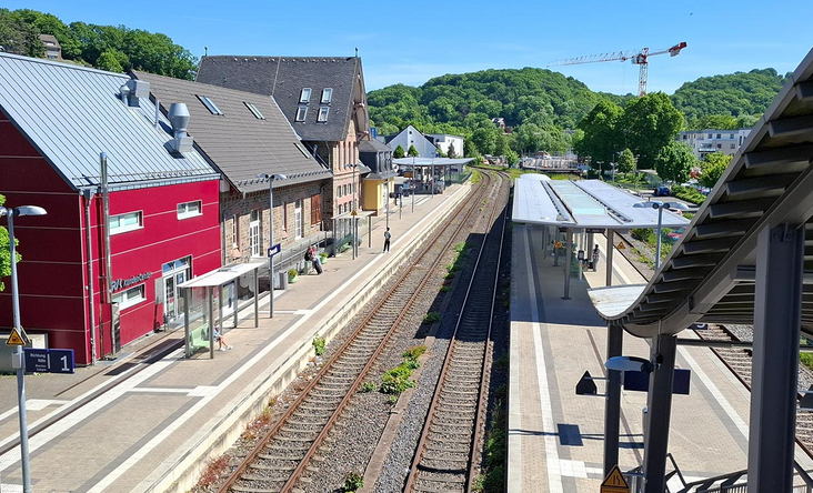 Bahnhof Overath aus der Vogelperspektive mit modernisierten Bahnsteigen, historischen Gebäuden und grünen Hügeln im Hintergrund, sonniger Sommertag