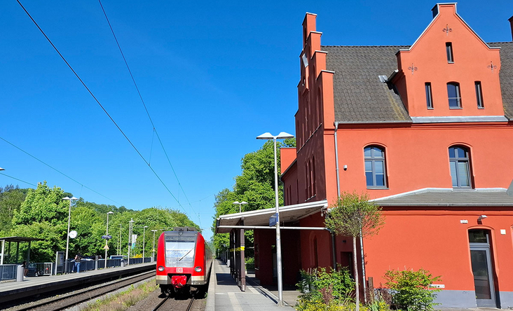 Roter Bahnhof Schladern mit einfahrendem Regionalzug, sonniger Bahnsteig, Oberleitungen und blauer Himmel