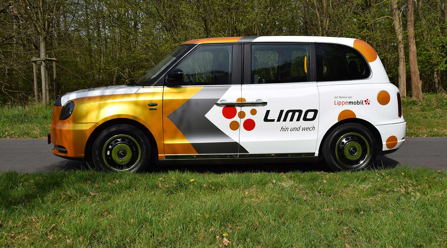 Ein On-Demand-Auto mit bunten Aufklebern und dem Schriftzug Limo steht auf einer Straße an einer Wiese.