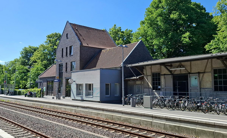 Bahnhof Billerbeck mit Gleisen, modernisiertem Backstein‑Gebäude und Fahrradabstellbereich, sonniger Himmel und viel Grün