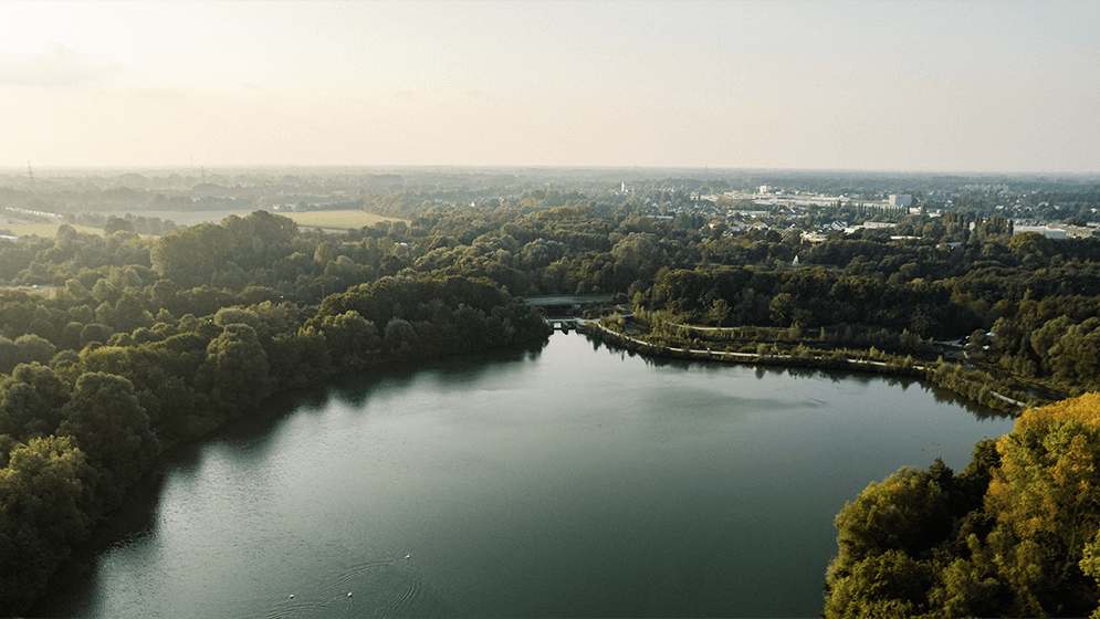 Zu sehen ist eine Luftaufnahme eines Sees umgeben von Wäldern. In der Ferne sieht man den Horizont. 