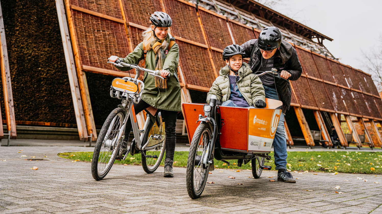 Eine Familie mit Vater, Mutter, Sohn fährt mit einem Leihrad und einem geliehenen Lastenrad durch die Natur. Der Sohn sitzt vorne in der Box des Lastenrads.