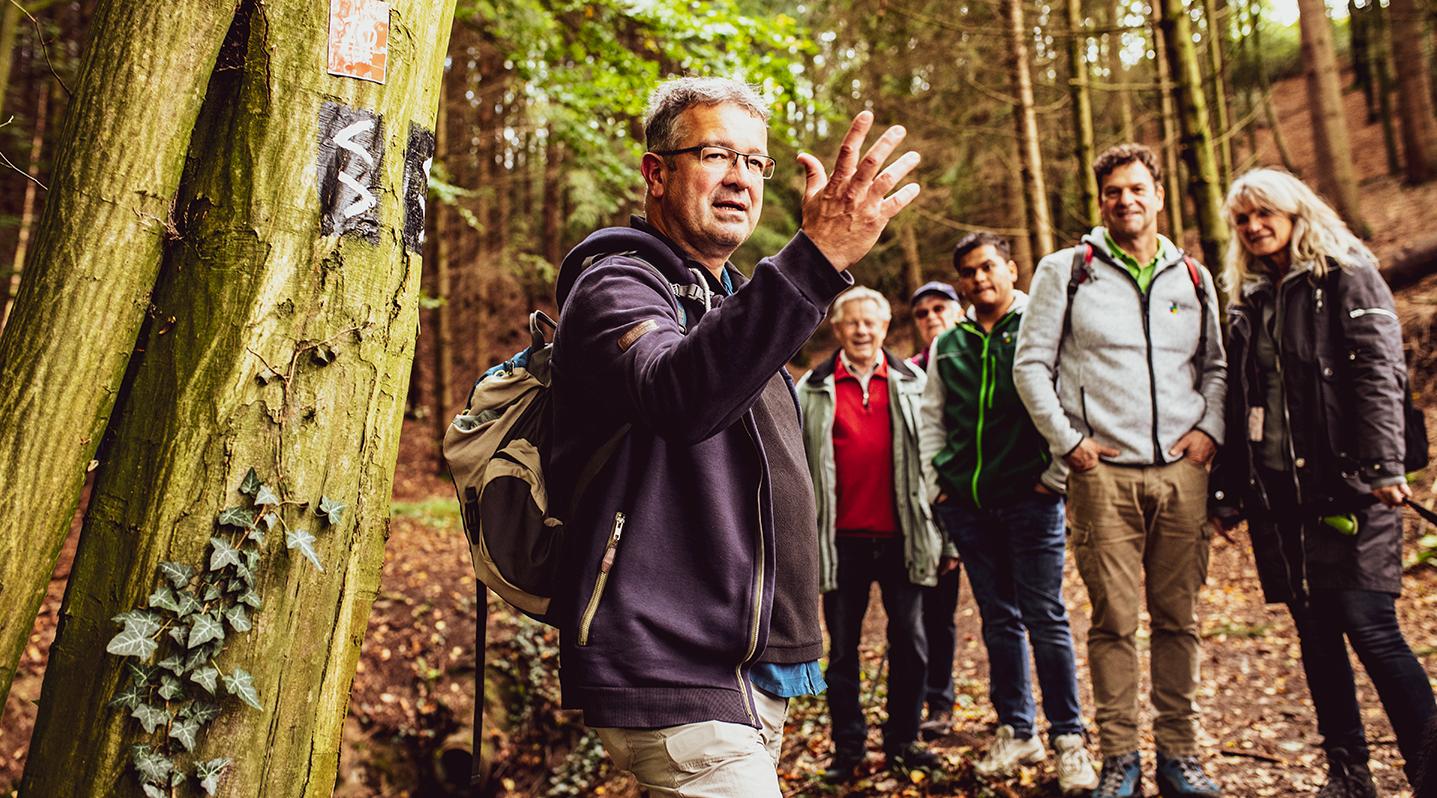Header Gewinnspiel Wanderbahnhof 2020 Manuel Andrack und eine Gruppe Wanderer steht im herbstlichen Wald, während Manuel Andrack etwas erklärt.
